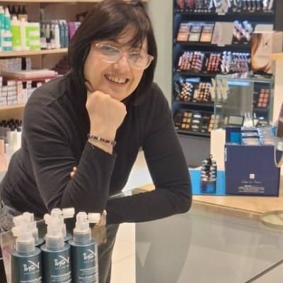 Persona sorridente appoggiata a un bancone in un negozio di cosmetici, con flaconi esposti in primo piano.