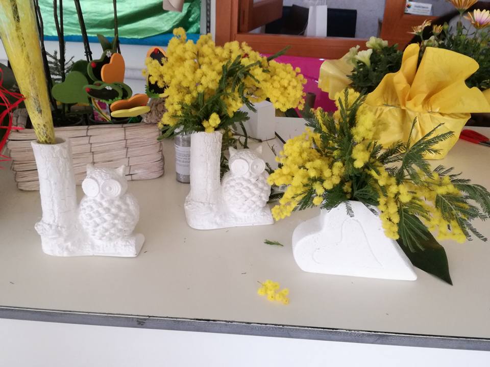 Vasi bianchi a forma di gufo con fiori di mimosa su una superficie bianca, con altre decorazioni floreali sullo sfondo.