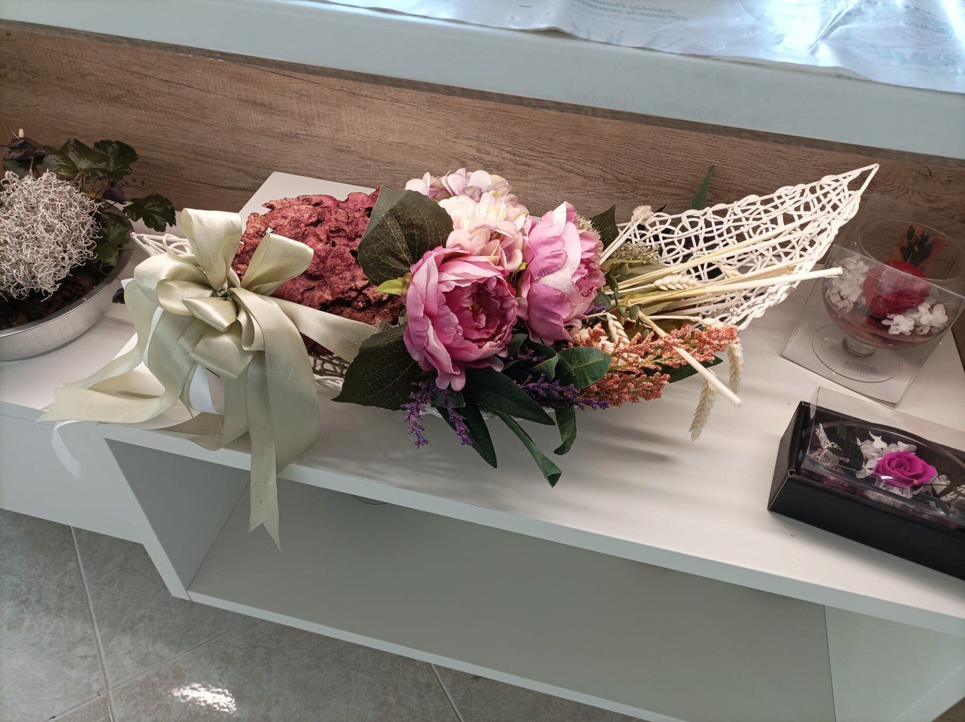 Composizione floreale con fiori rosa, nastri e dettagli decorativi su una mensola bianca.