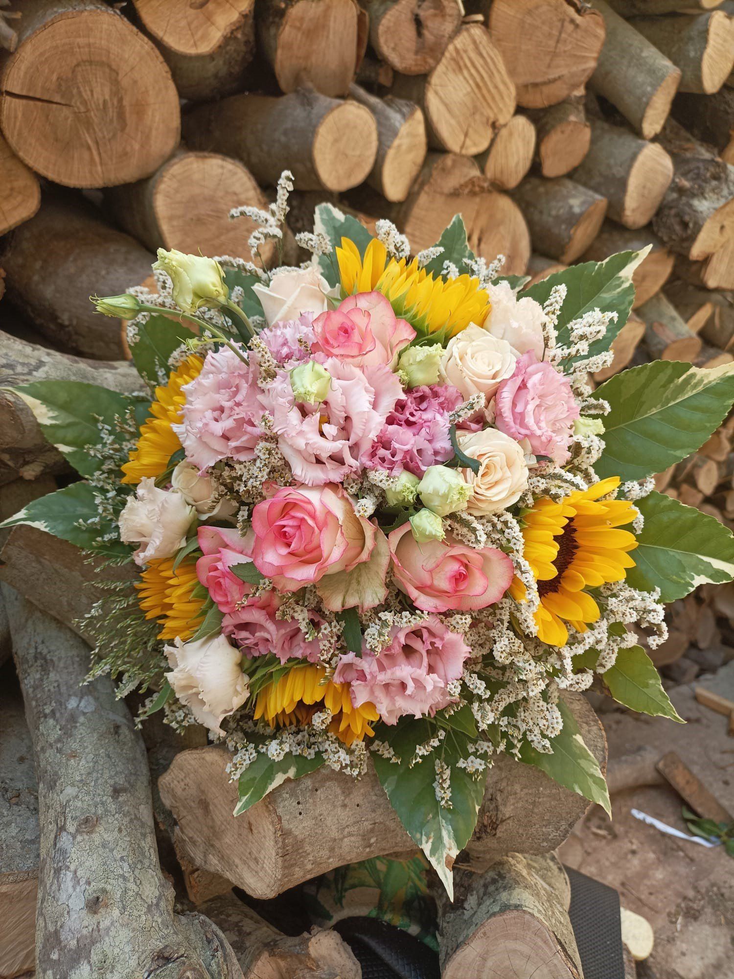 Mazzo di fiori rosa, gialli e bianchi con fogliame verde, davanti a una catasta di legna.