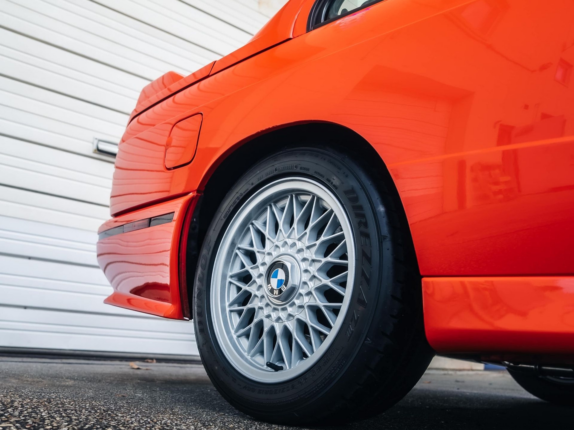 Bright orange BMW E30 M3, rear quarter view, silver wheel, parked in front of a white garage door.