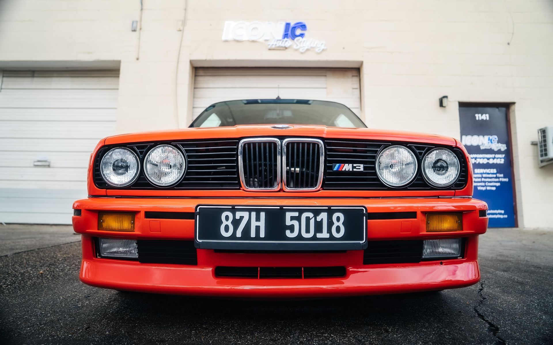 Bright orange BMW E30 M3, front view, parked in front of a building with a closed garage door.