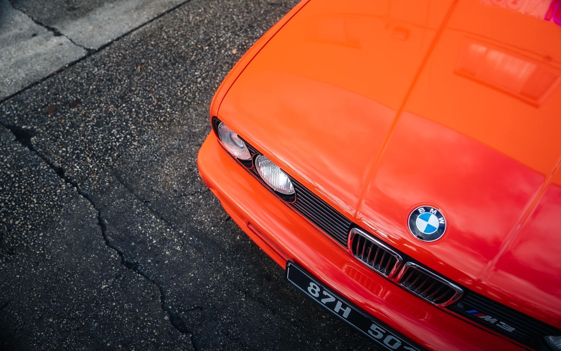 Close-up, high-angle view of the front corner of a bright orange BMW parked on asphalt.