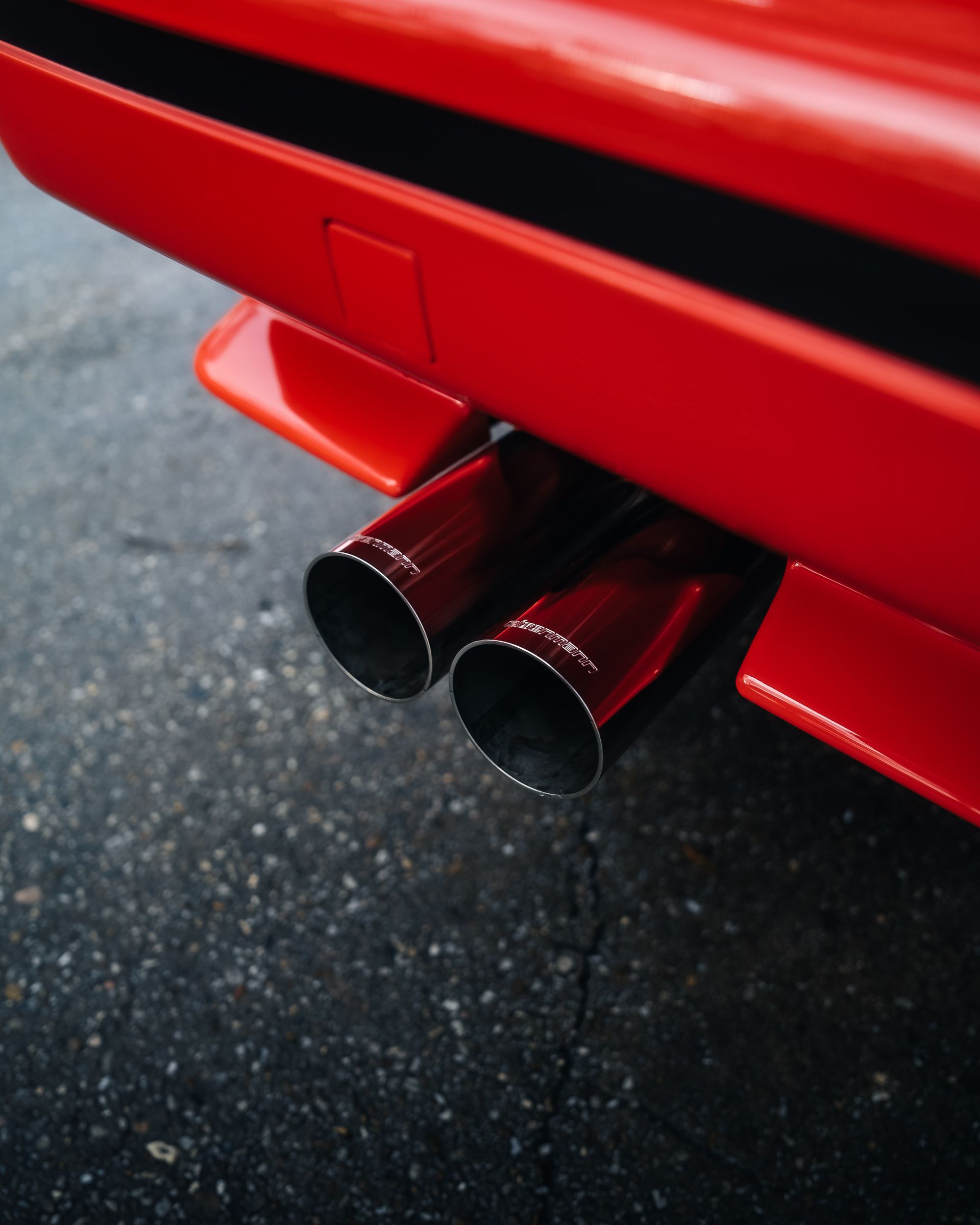 A close-up of a bright red car bumper with two shiny, circular exhaust pipes, positioned over a dark asphalt surface.