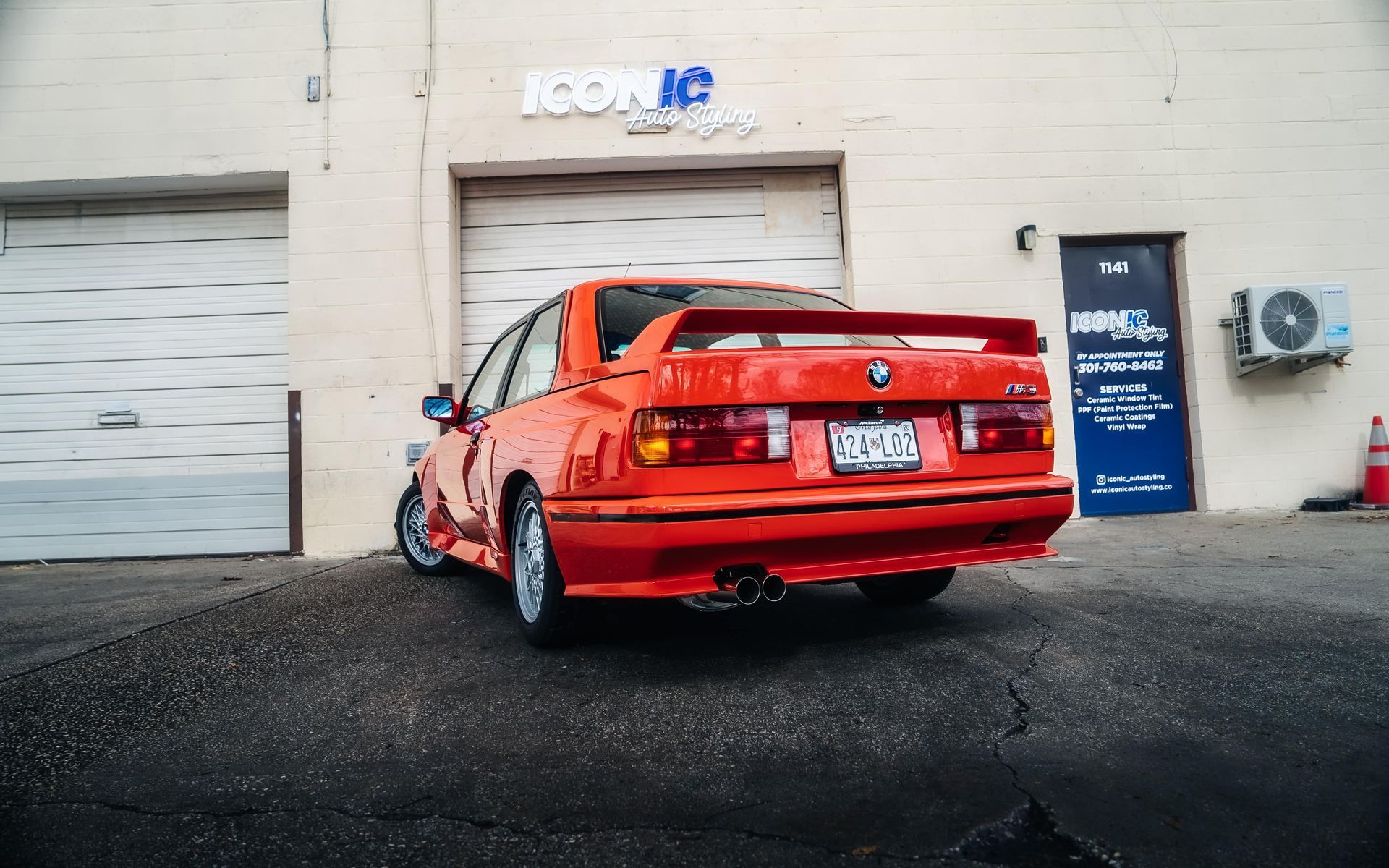 Orange BMW E30 M3 parked outside a garage, with a rear view showing the spoiler and dual exhaust.
