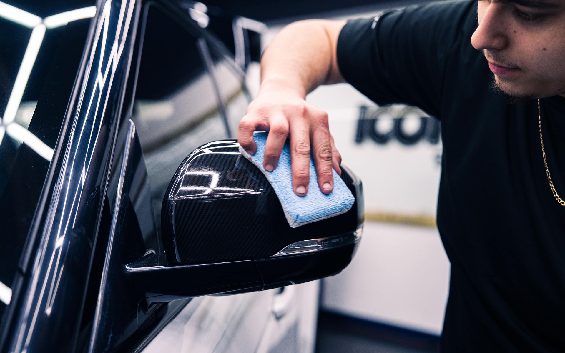 Person polishing a black car mirror with a blue cloth, inside a garage.