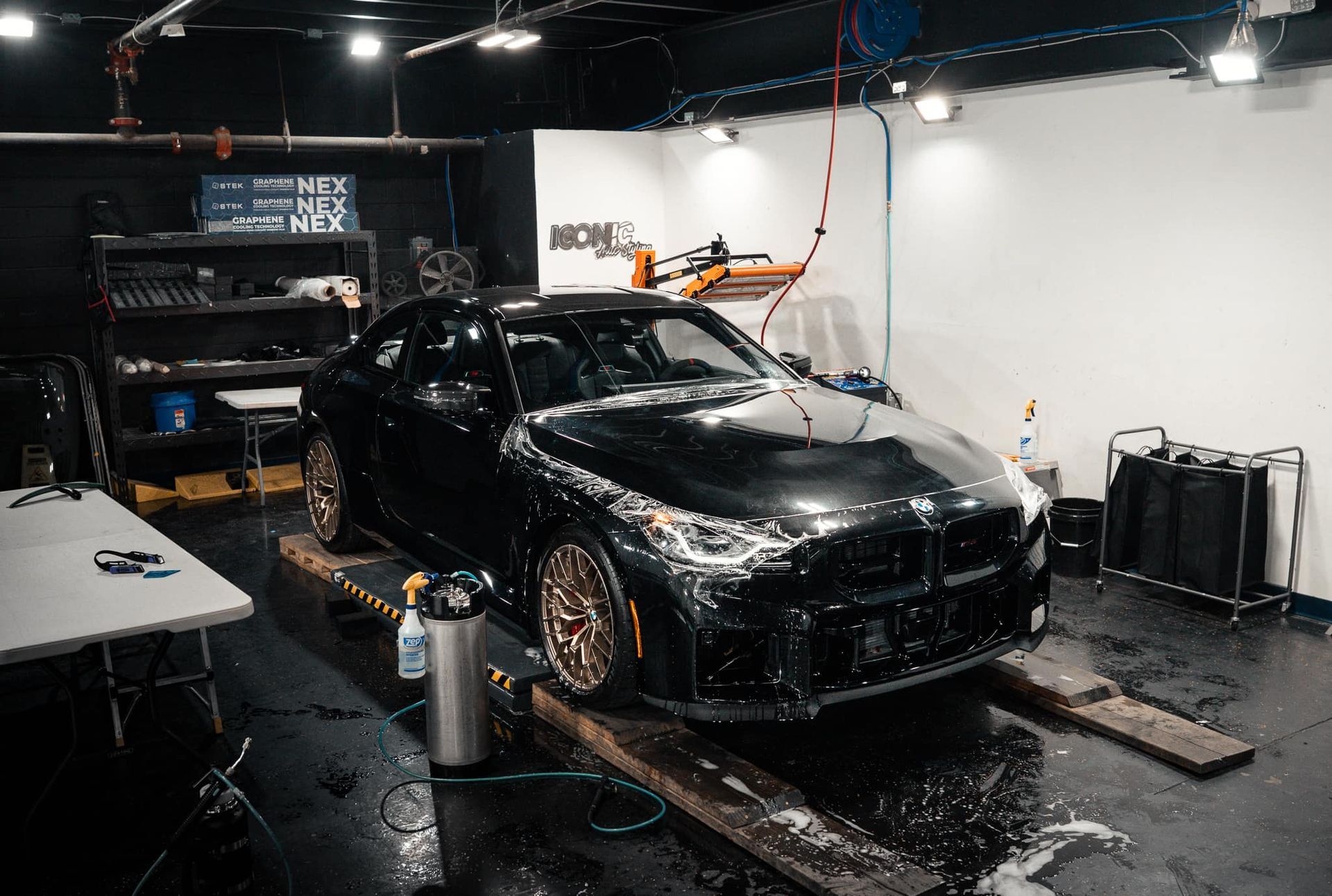 Black BMW being washed inside a garage; car on wooden ramps, covered in soap.