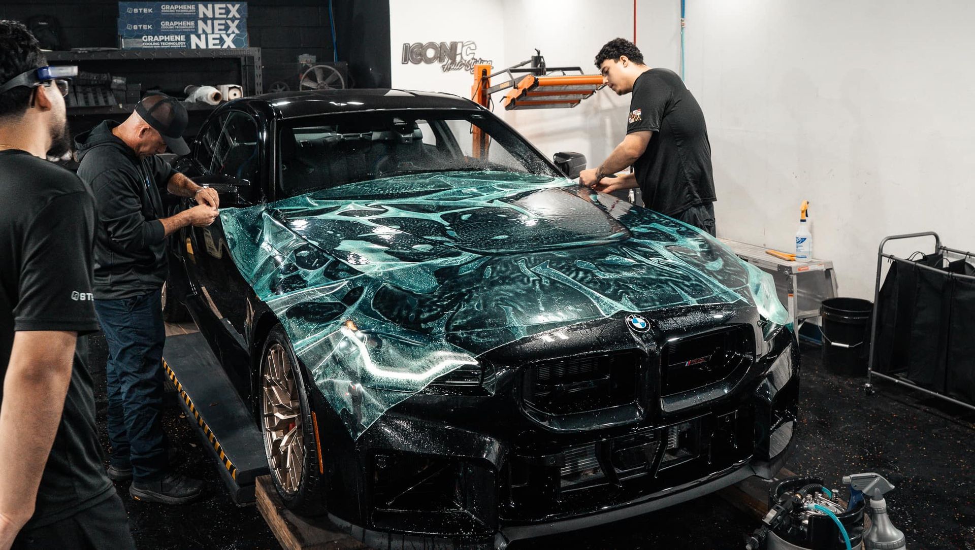 Three people wrapping a black BMW with a blue and white patterned vinyl wrap in a garage.