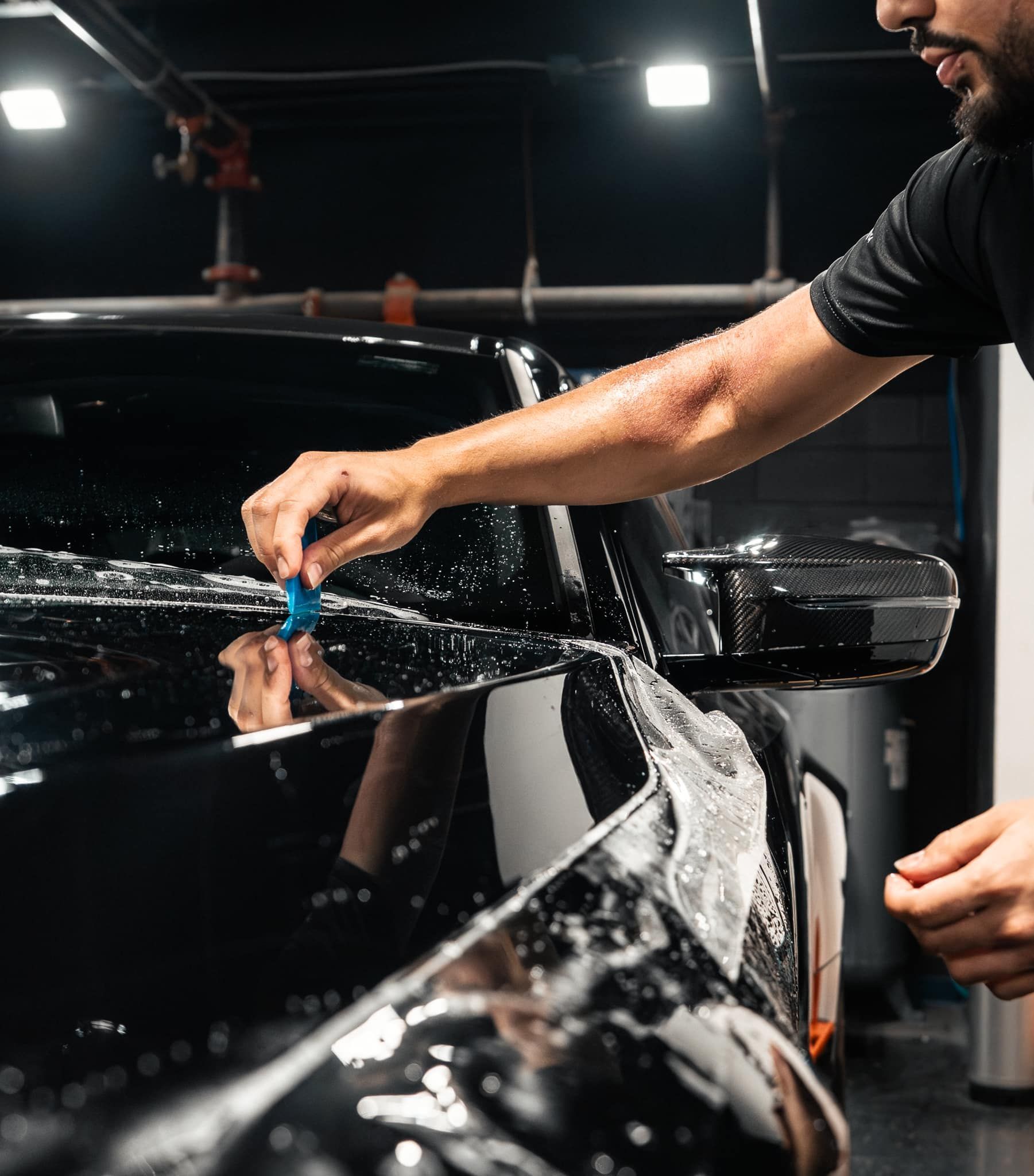 A person applying protective film to a black car, using a squeegee and spray bottle in a garage.