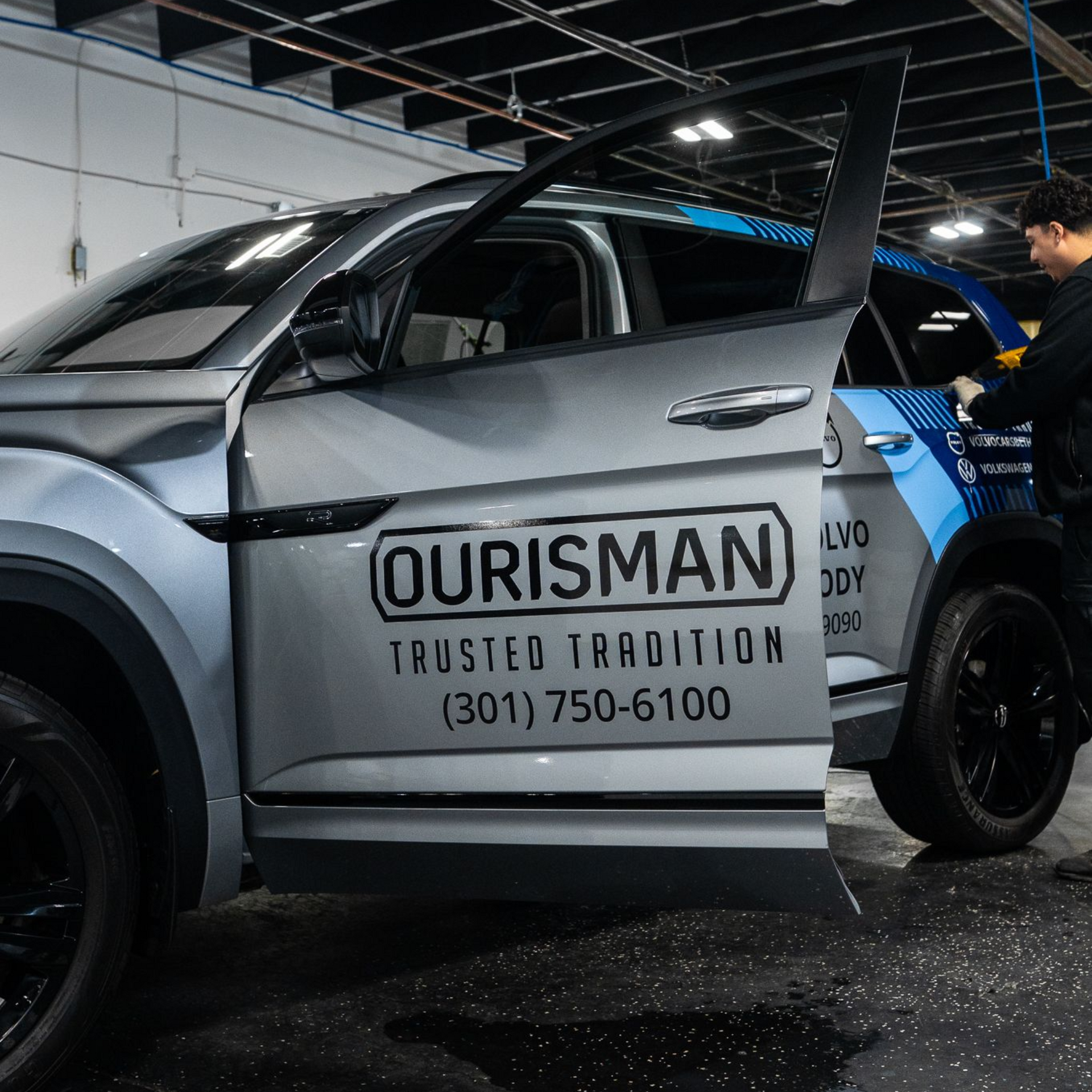 A technician works on a silver Ourisman-branded SUV with an open door inside an automotive shop.
