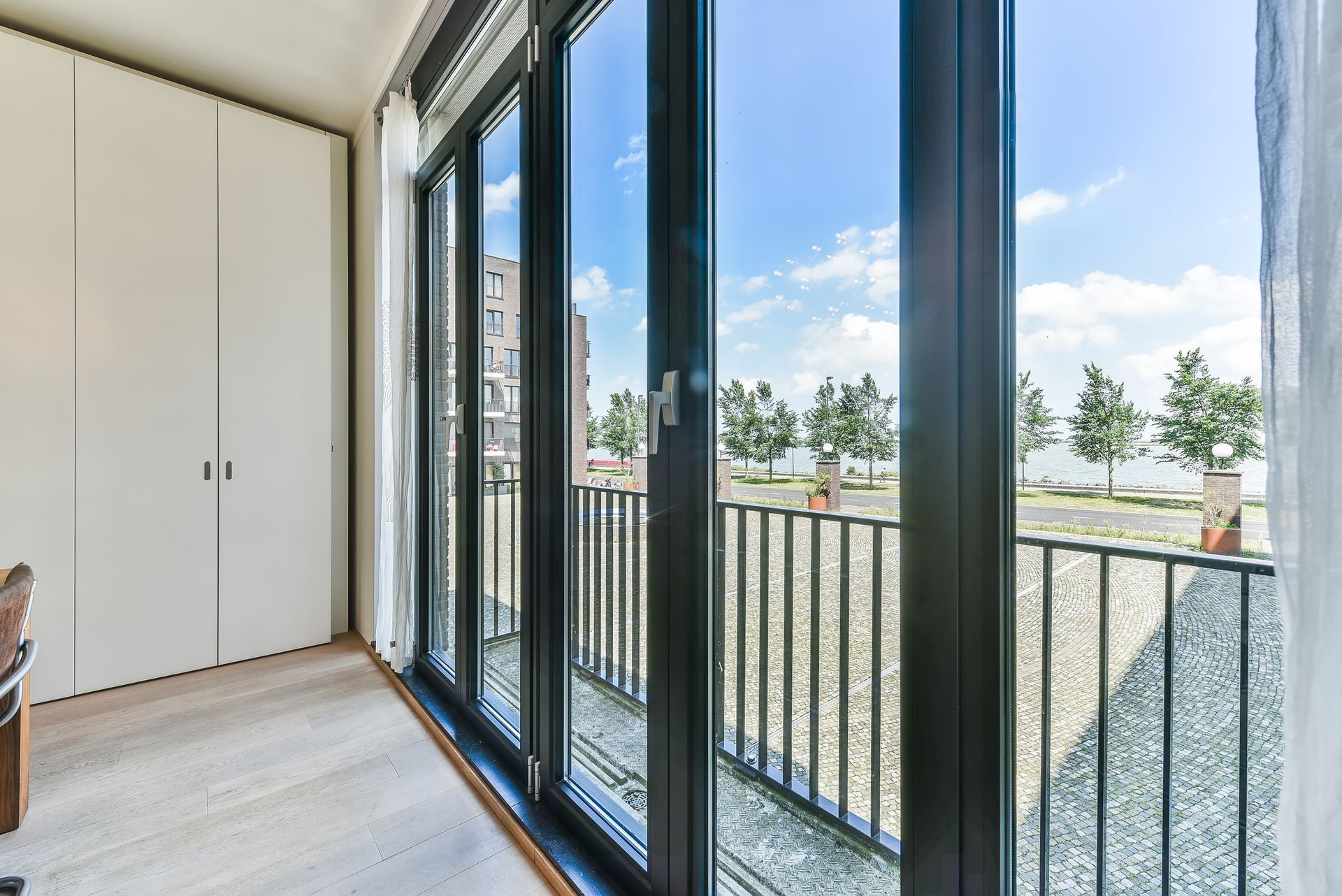Interior view with a large window and a balcony overlooking a brick paved area, trees, and buildings against a blue sky.