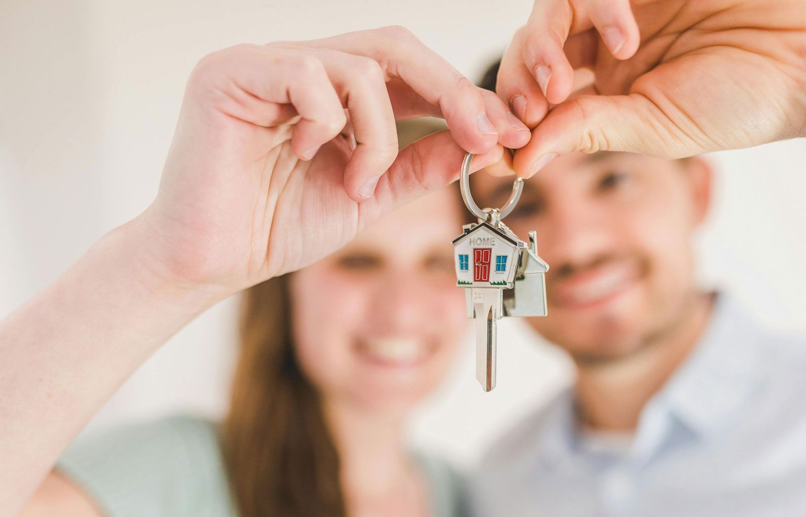 Hands holding a house key with a small house charm, with a smiling couple blurred in the background.