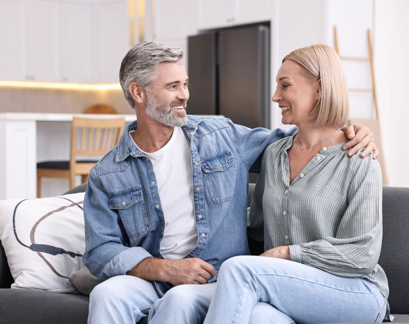 Two people smiling on a couch in a bright living room, chatting closely with arms around each other.
