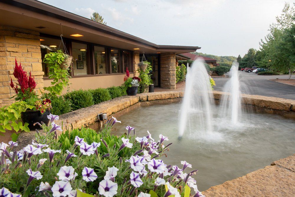 Fountain with two water jets in front of a tan building with hanging flower baskets and a road.