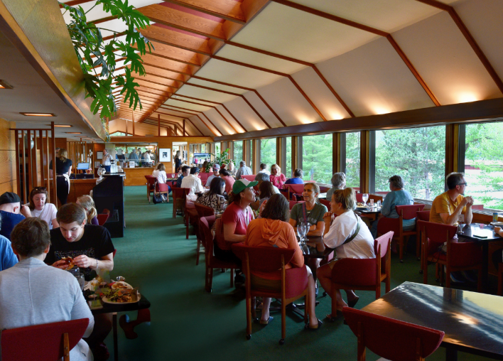 Restaurant interior with diners seated at tables. Large windows with a view. Green carpet, red chairs.