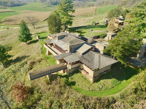 Aerial view of a complex, low-lying building with multiple connected sections, set in a grassy landscape with trees.