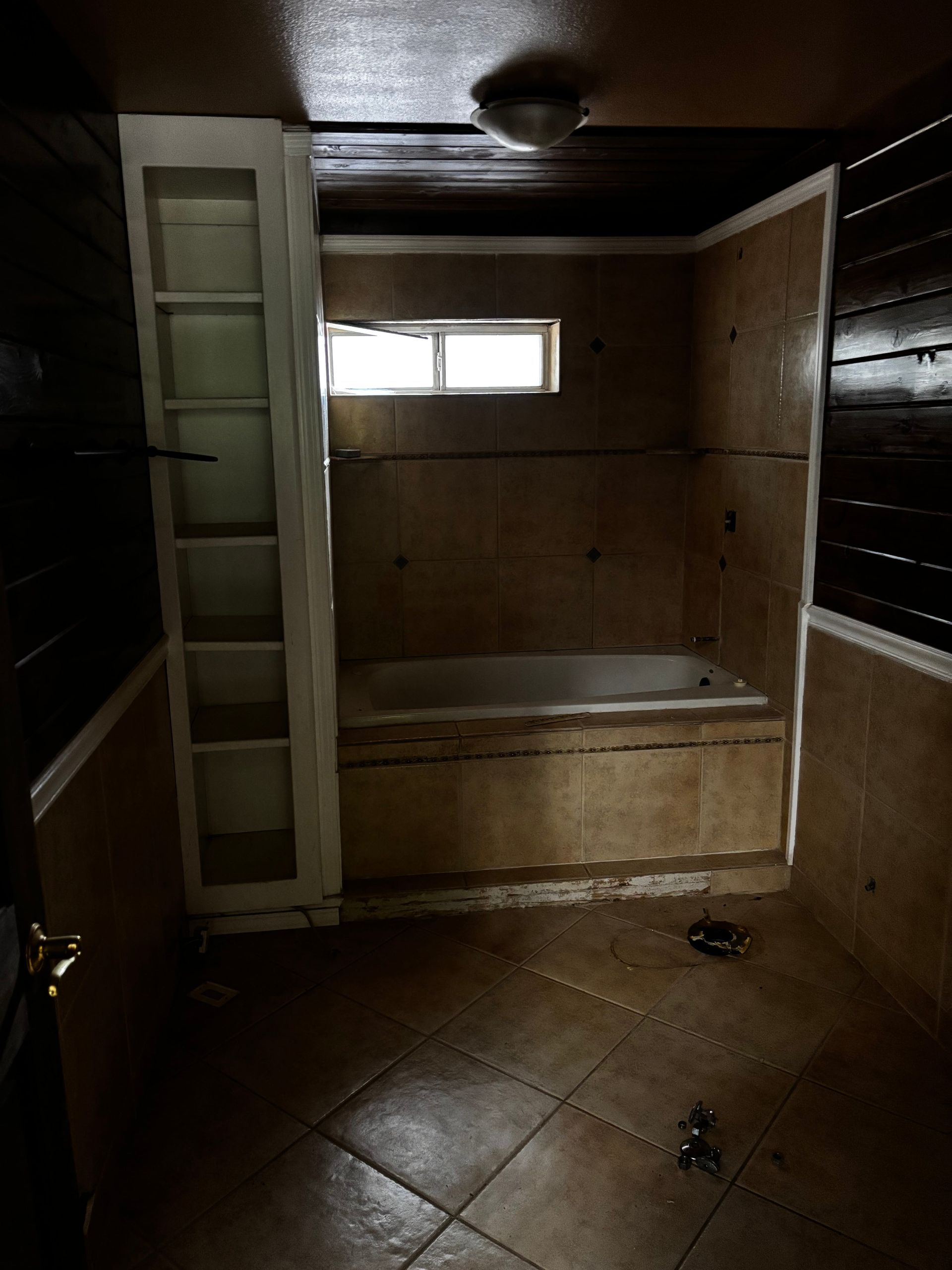 Bathroom with tan tile, built-in tub, and a small window. Dark wood paneling on walls. White shelf unit.