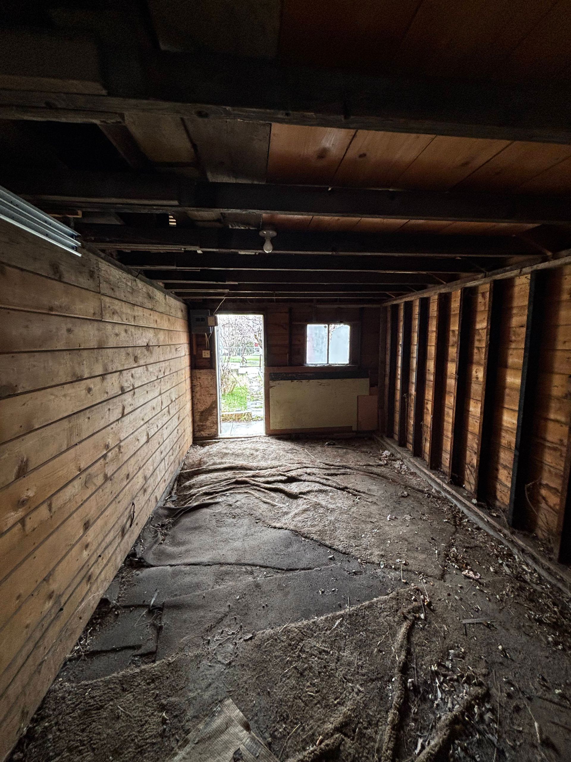 Interior view of a dilapidated wooden structure. Walls are partially stripped; debris covers the floor. Door and window visible.