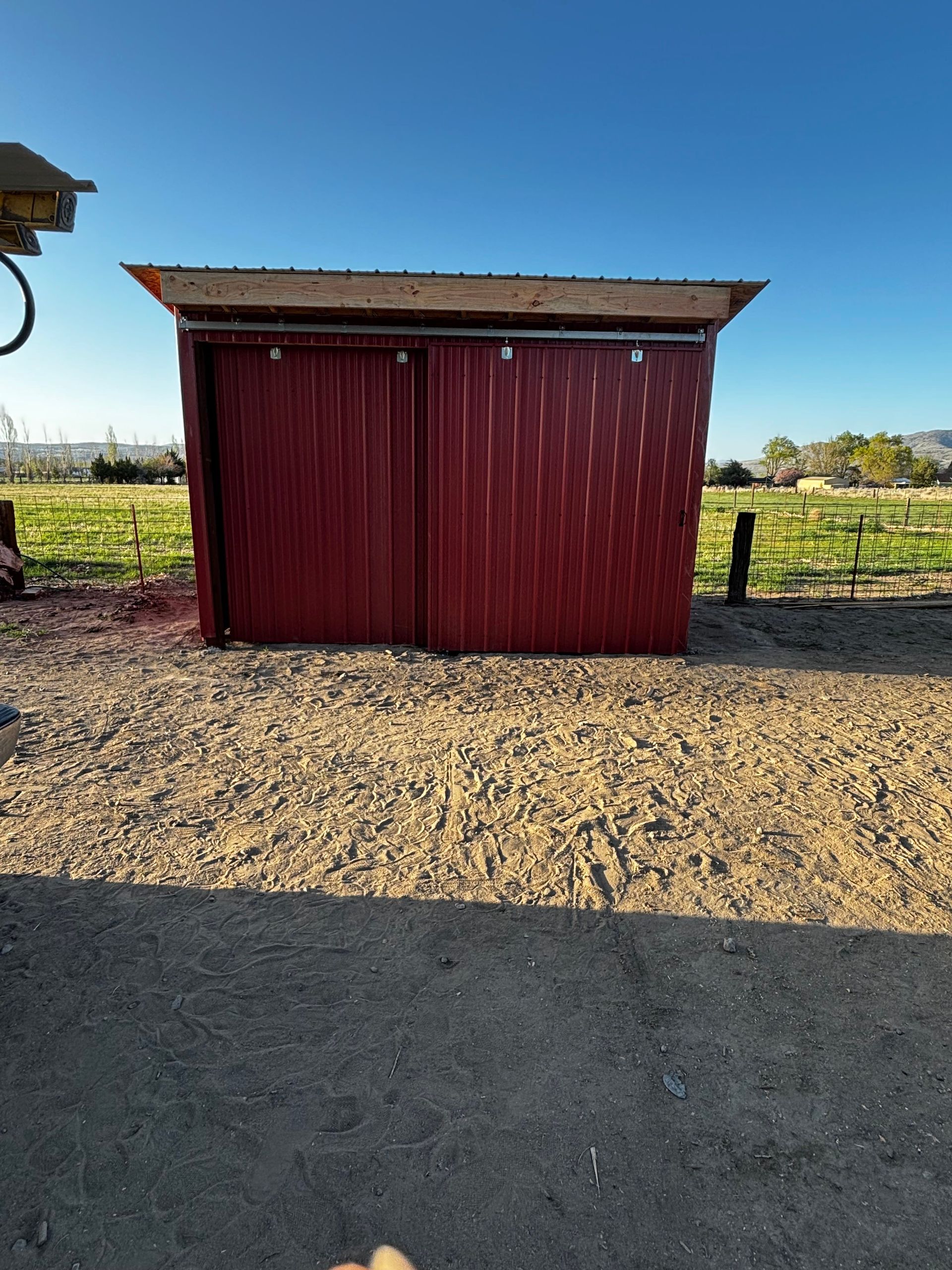Red metal shed with sliding doors on gravel, under a blue sky.