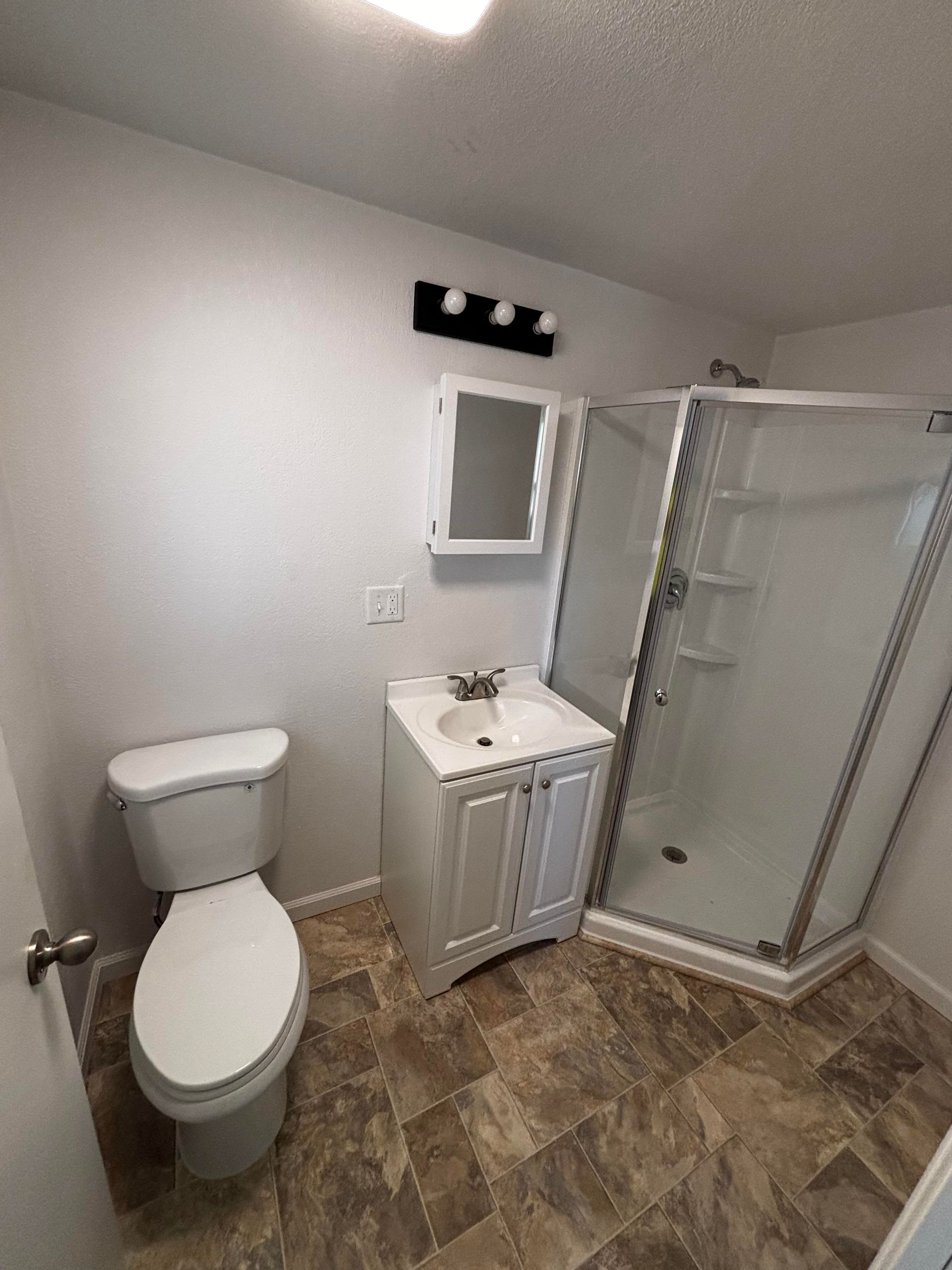 Bathroom with a white toilet, vanity, and corner shower. Brown patterned flooring.