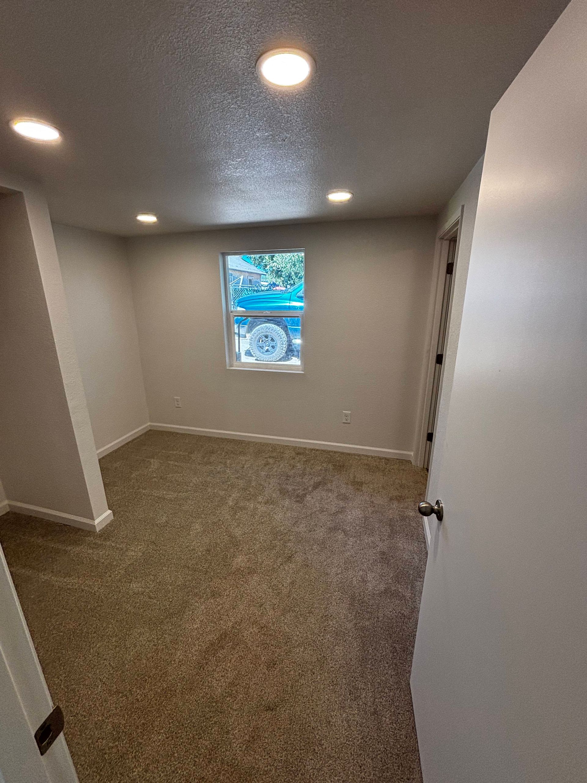 Empty bedroom with tan carpet, white walls, three ceiling lights, and a small window.