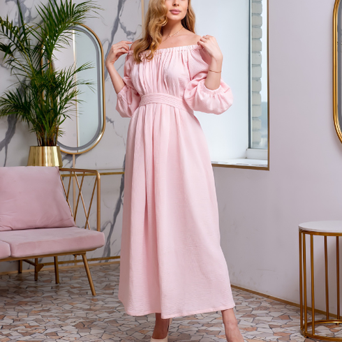 Woman in a light pink off-shoulder dress posing in a bright room with a plant, chair, and round mirror.