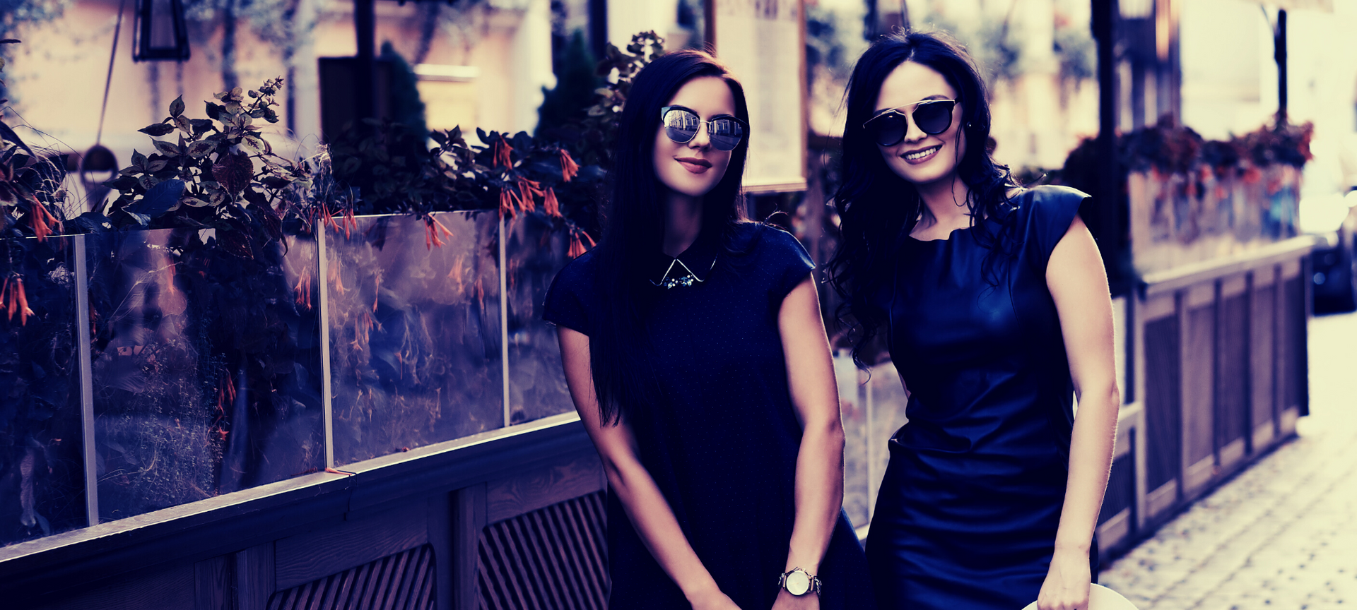 Two women in black dresses and sunglasses standing by a flower-lined wall outdoors.