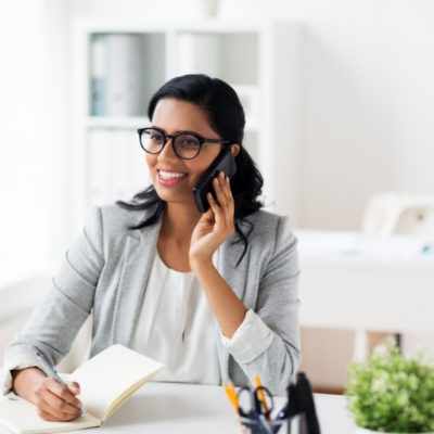 Woman with glasses smiling while talking on a phone at a desk, holding a notebook in a bright office
