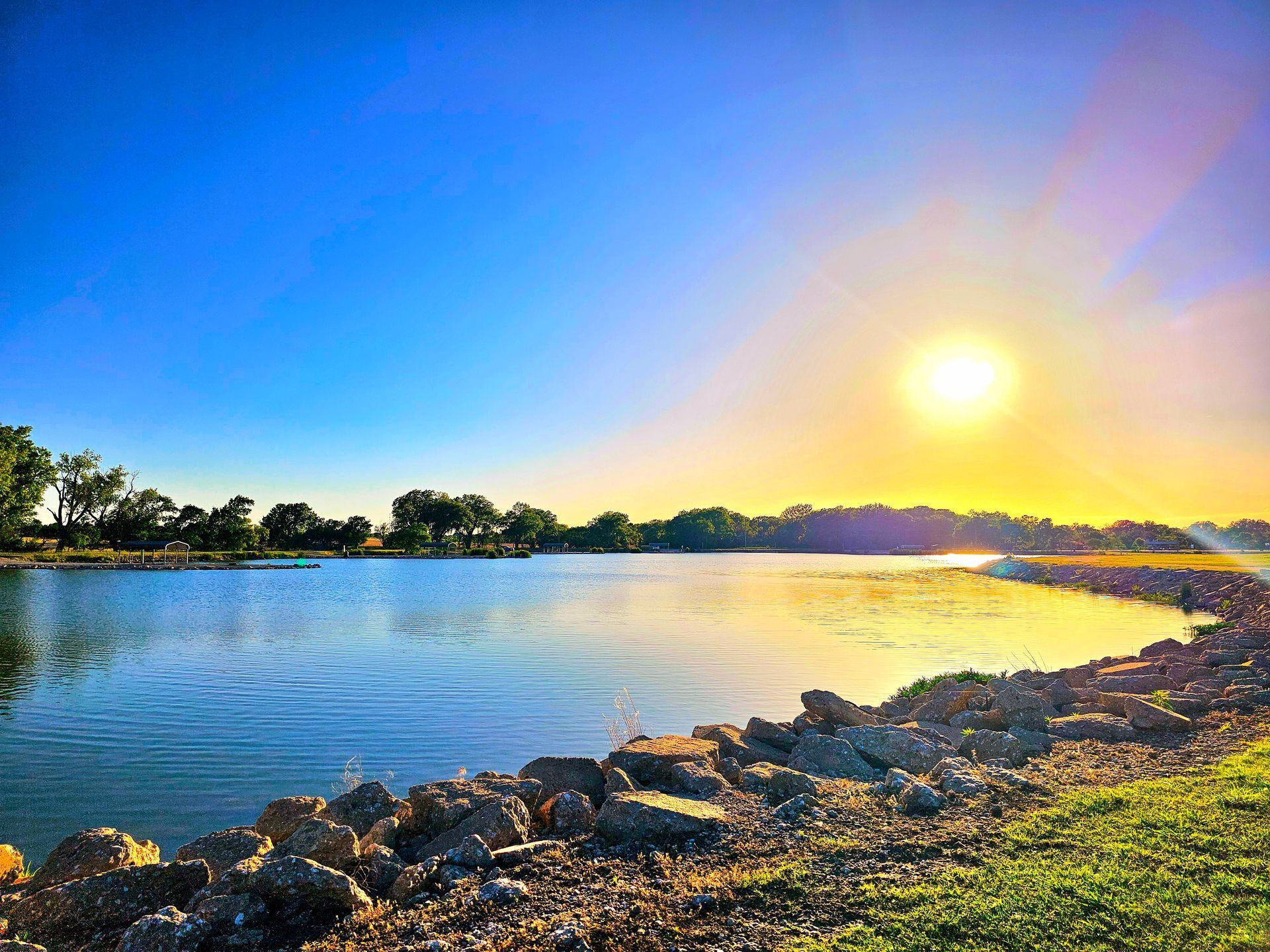 Sunset over a calm river with rocky shore, blue sky, and sun reflecting on the water