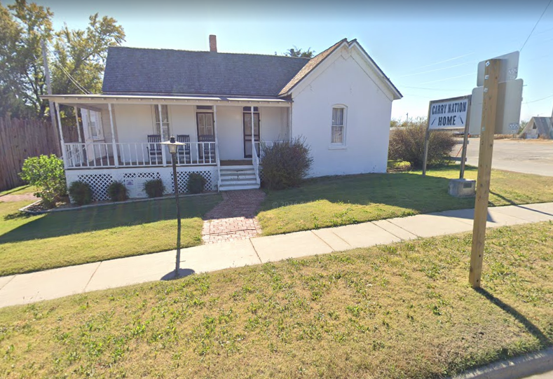 White house with front porch, small lawn, sidewalk, and a street sign on a sunny corner lot.
