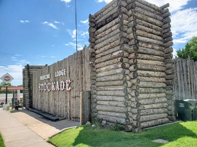 Wooden lodge sign reading βStockadeβ beside a rustic log-fence entrance under a blue sky