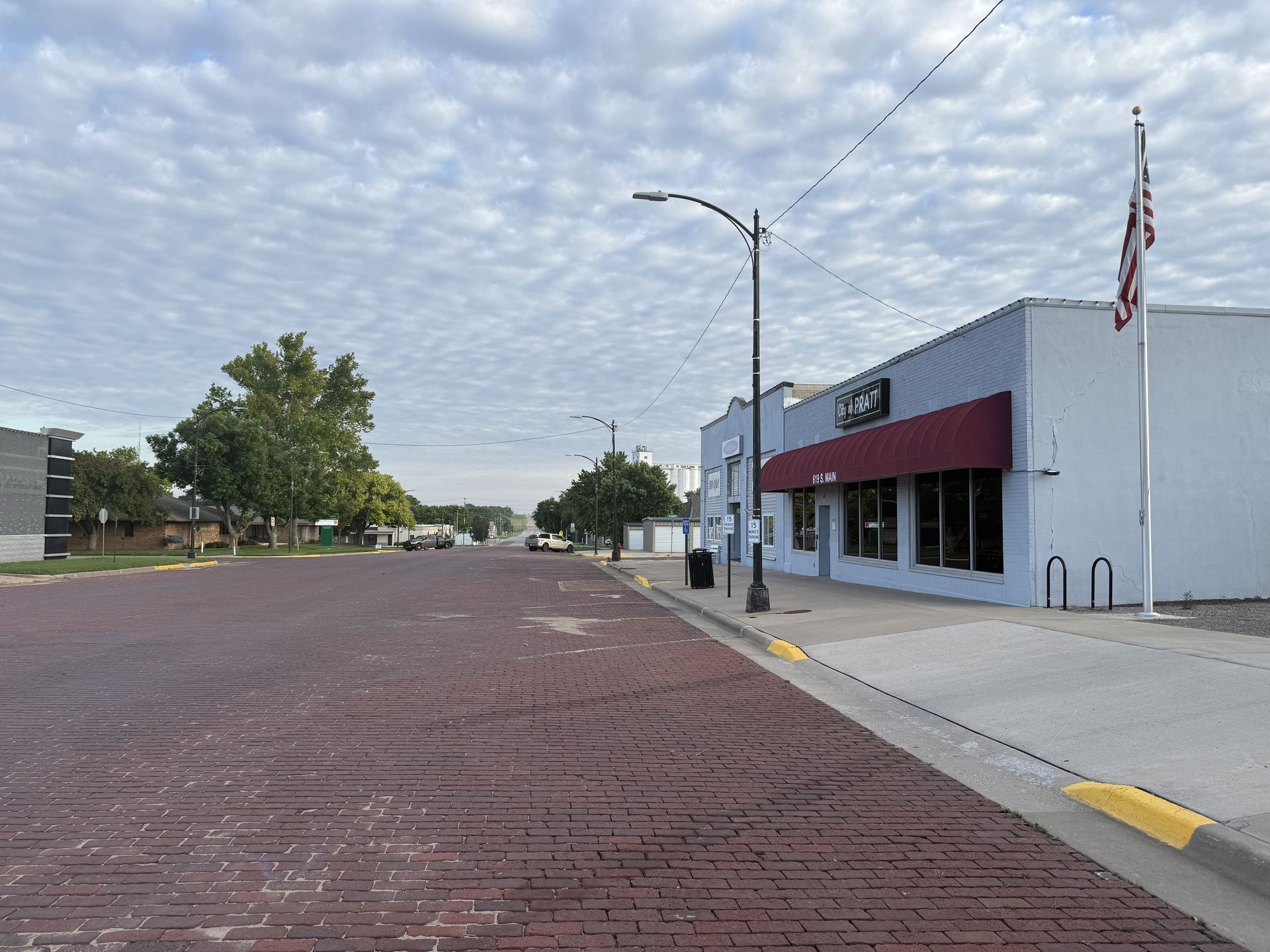 Quiet brick street with a small white storefront and an American flag under a cloudy sky.