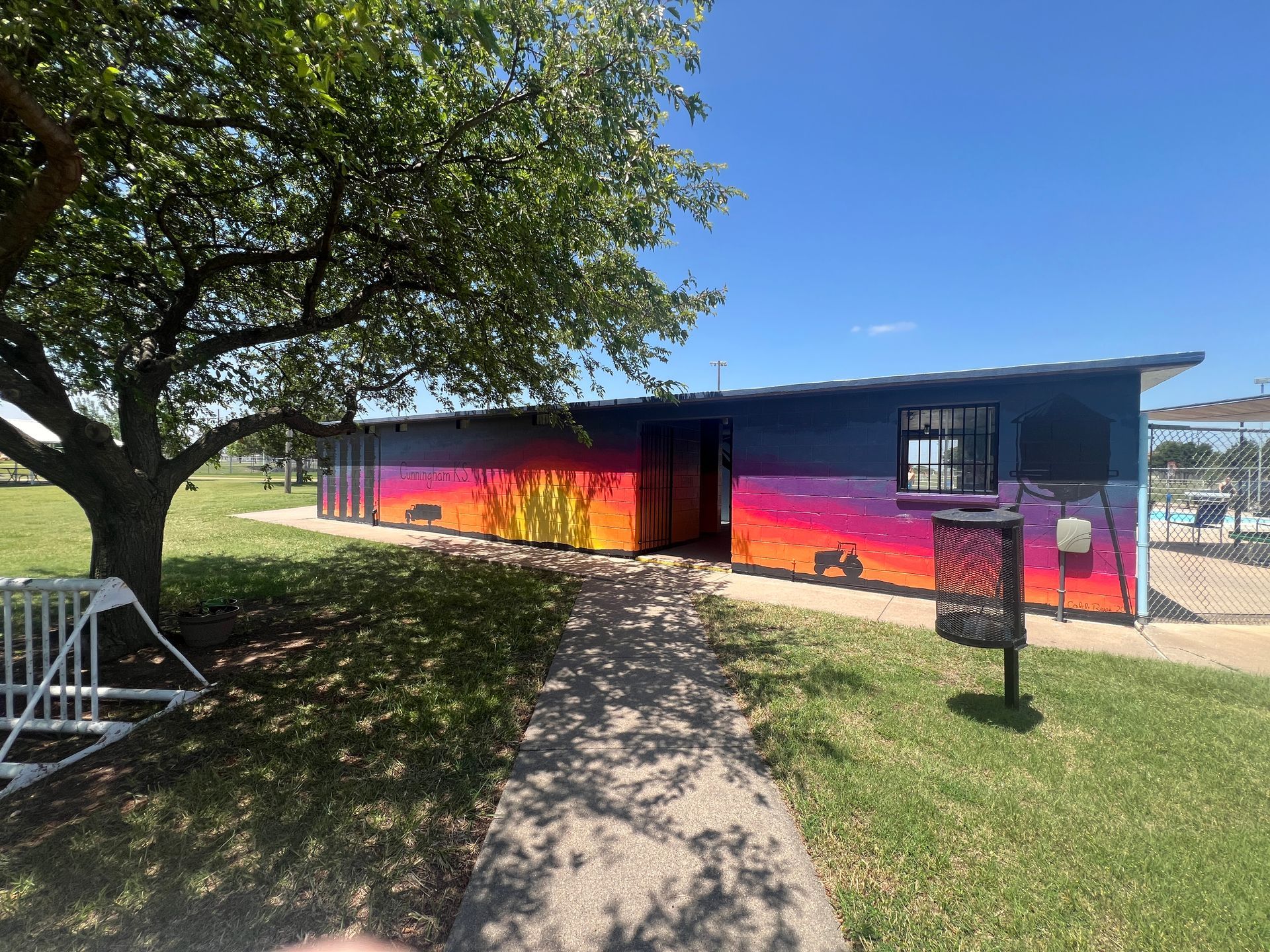 Colorful mural on a long building under a tree, beside a grassy path on a sunny day.