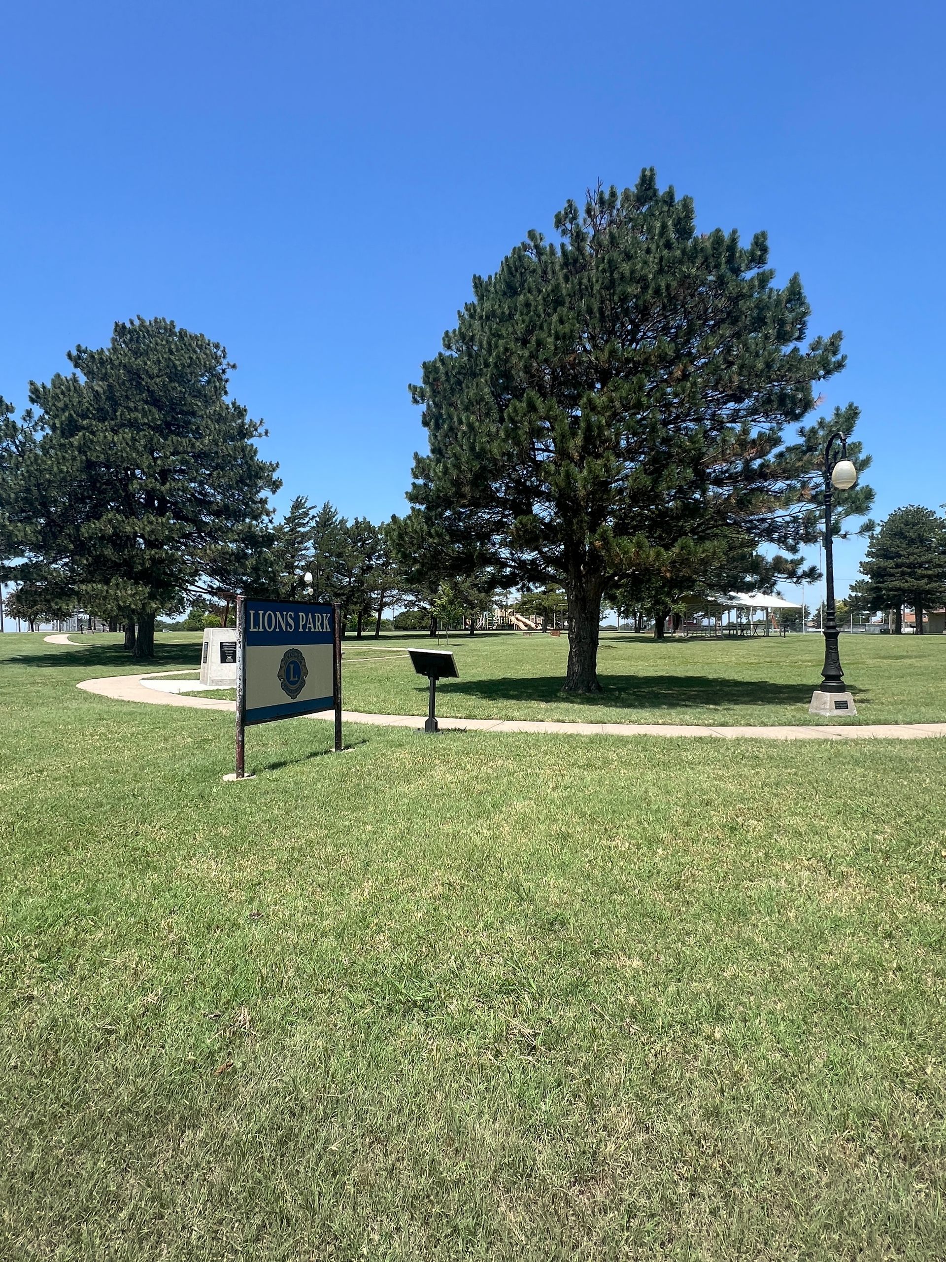 Sunny park with green grass, trees, and a small sign near a paved path.