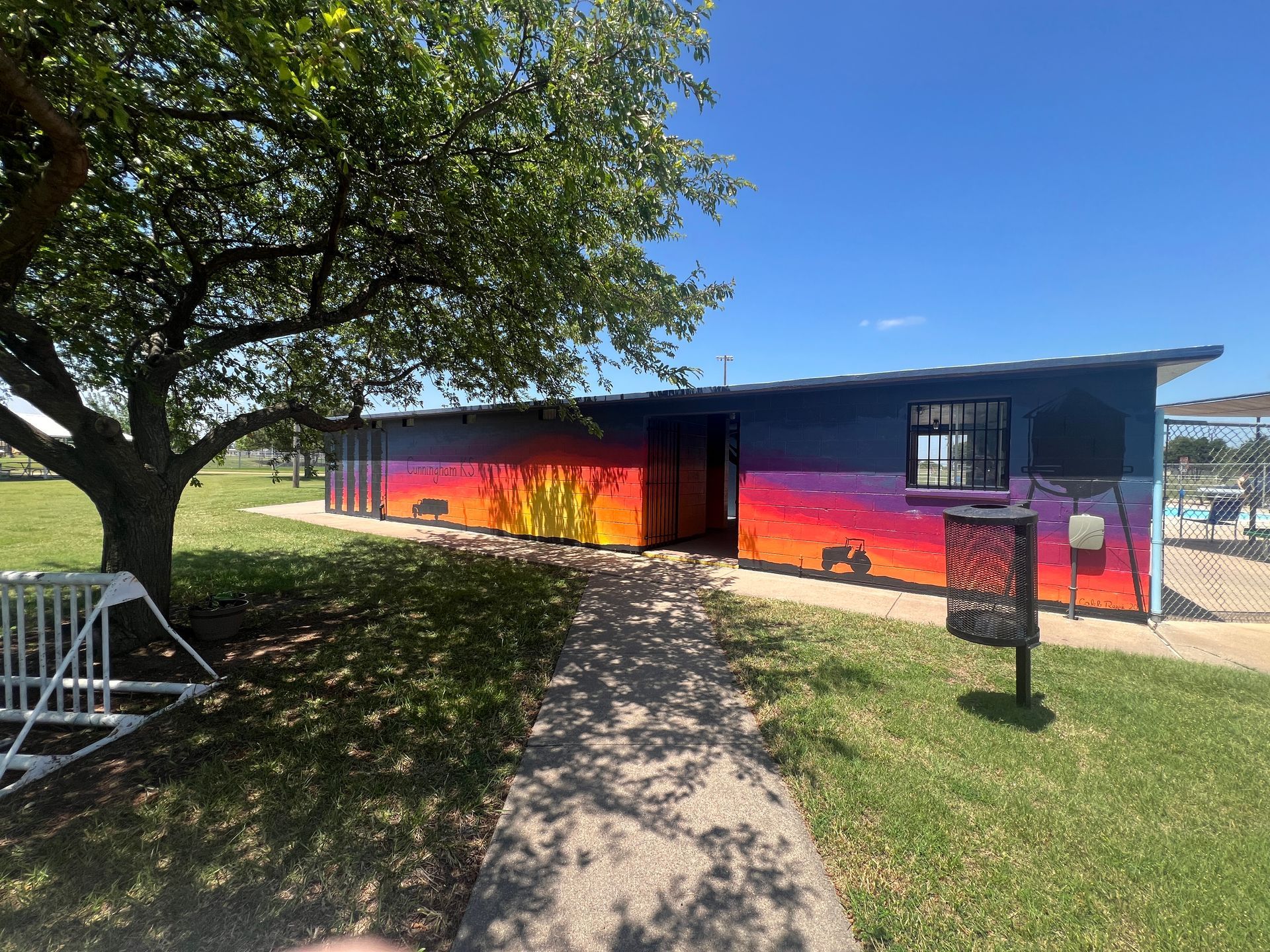 Long low building with colorful sunset mural, tree shade, and blue sky on a grassy lawn