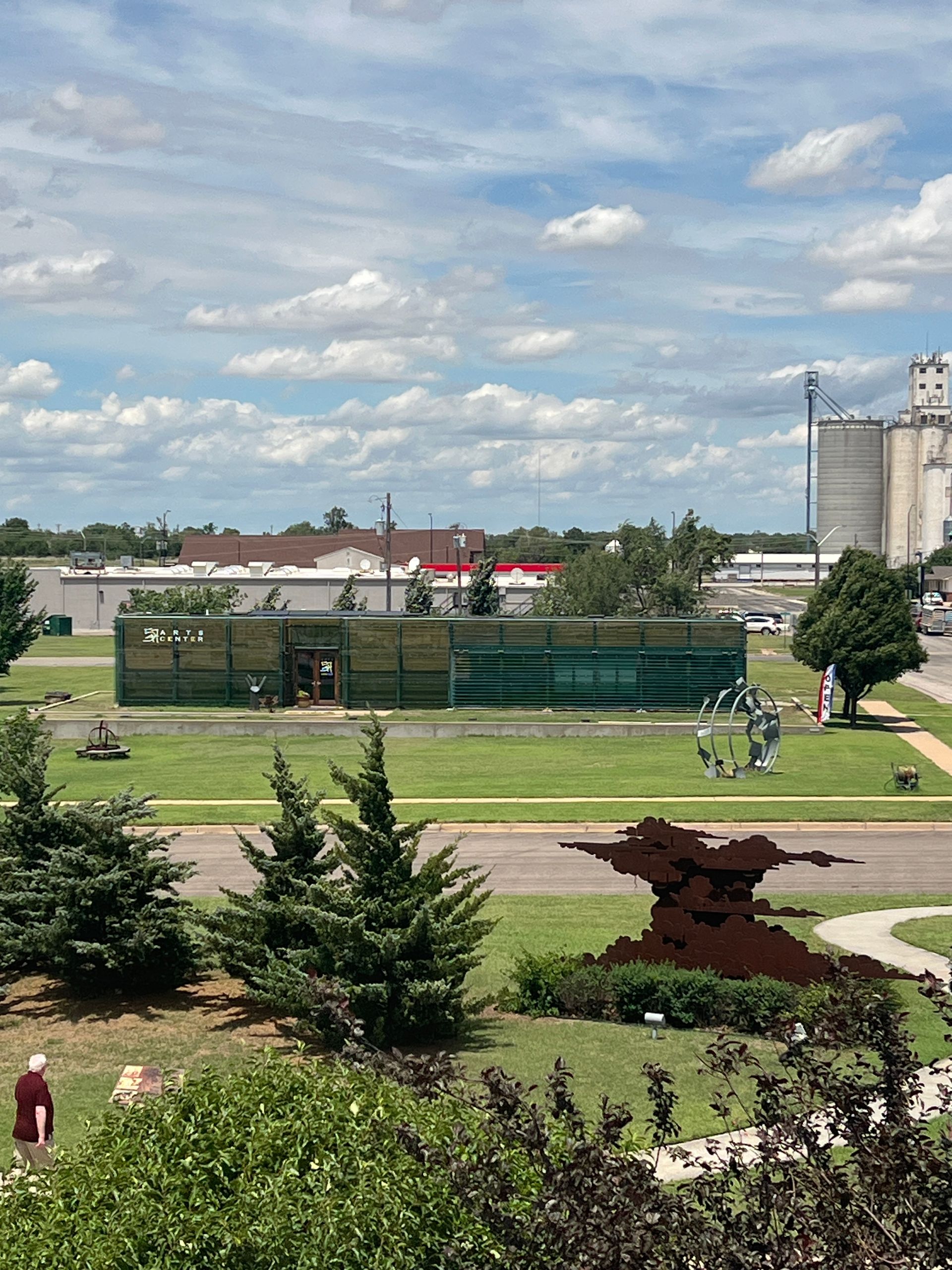 Green park with trees and pathways, a fenced sports court, and silos under a cloudy sky.
