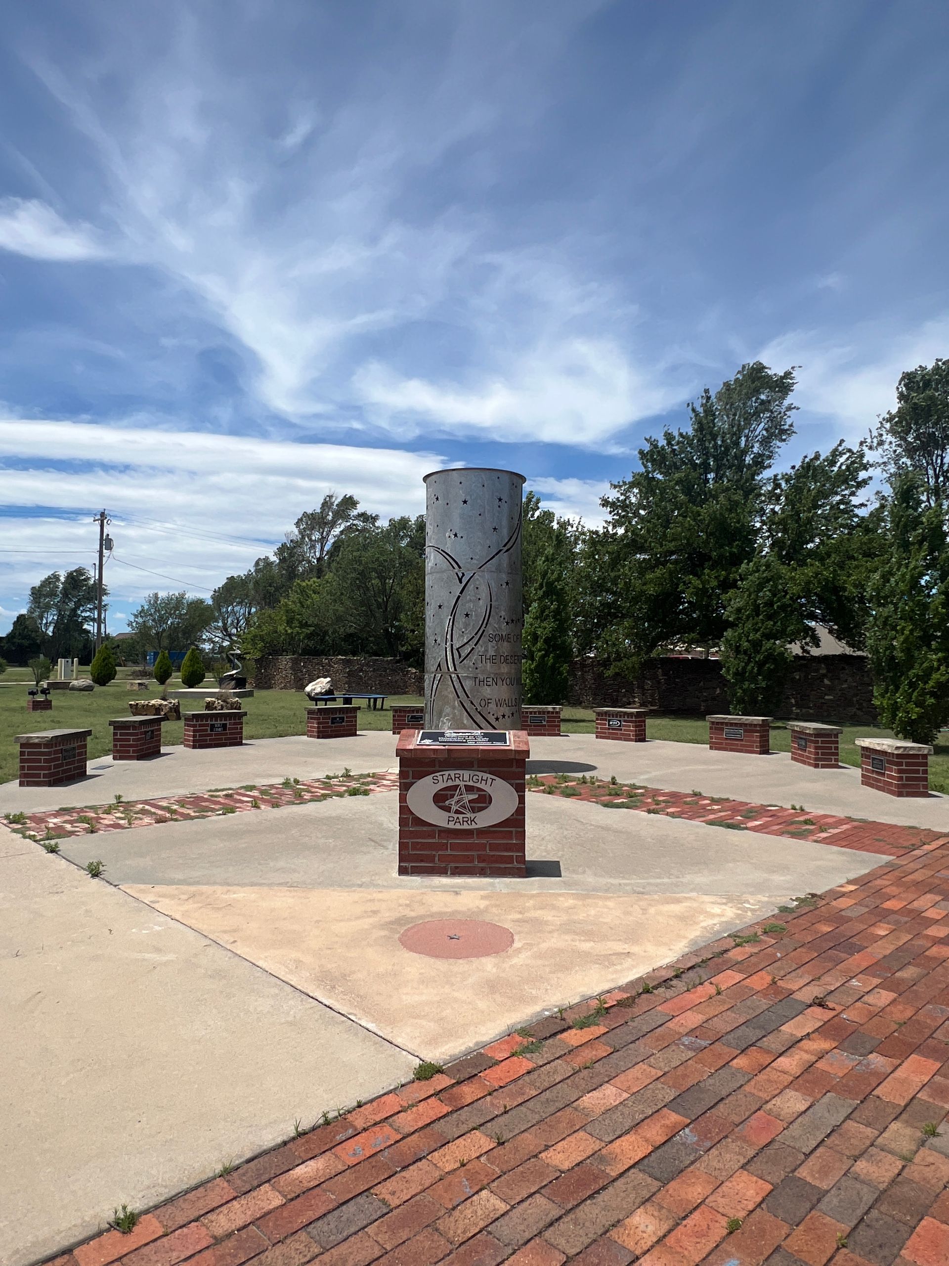 Outdoor memorial with a concrete pillar, brick walkway, benches, and a blue sky with clouds