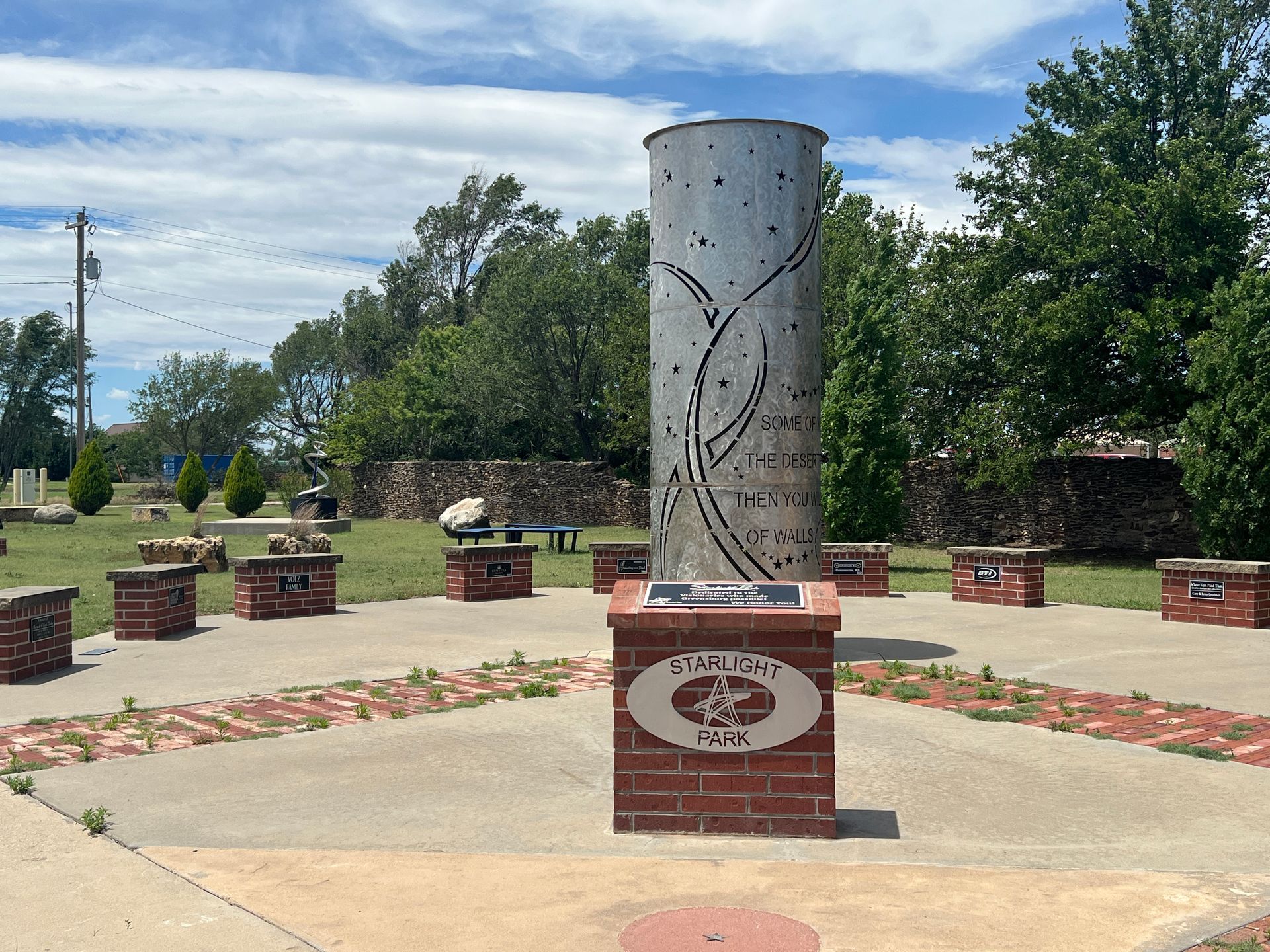 Outdoor memorial column in a brick plaza with benches and trees under a partly cloudy sky