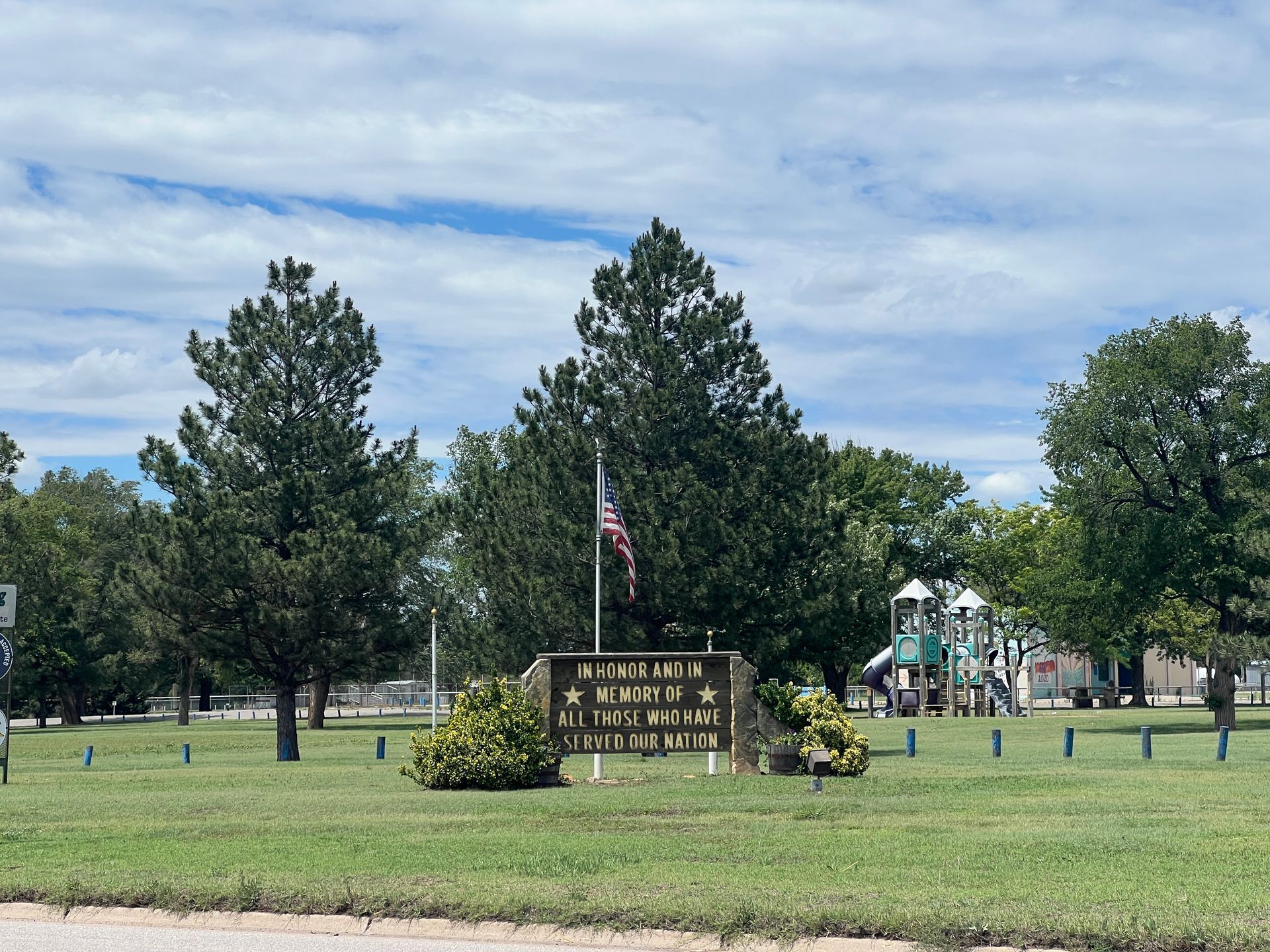 Memorial sign and U.S. flag in a grassy park with trees under a partly cloudy sky