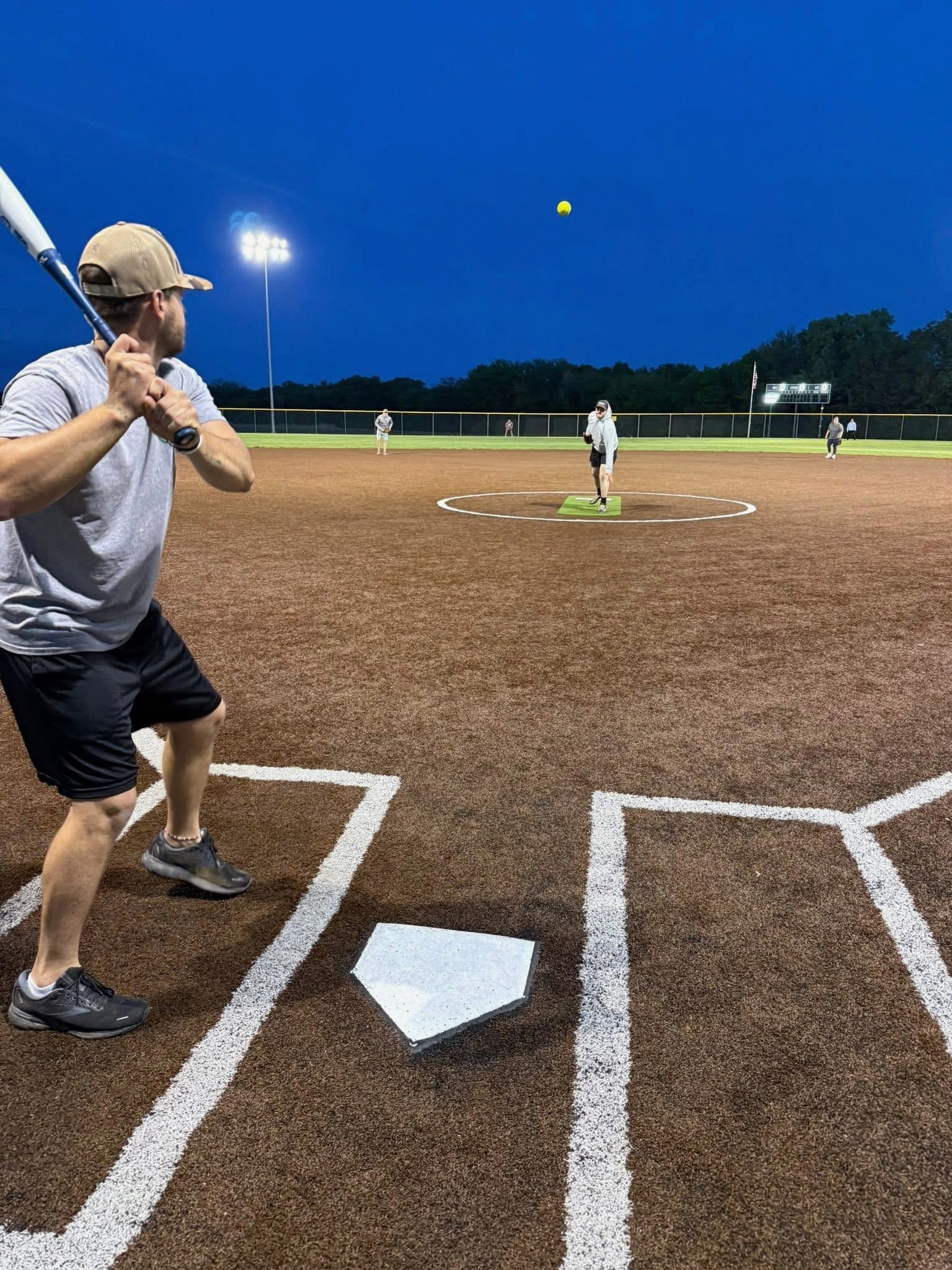 Batter at home plate under stadium lights, pitcher on mound, ball in midair during a nighttime baseball game