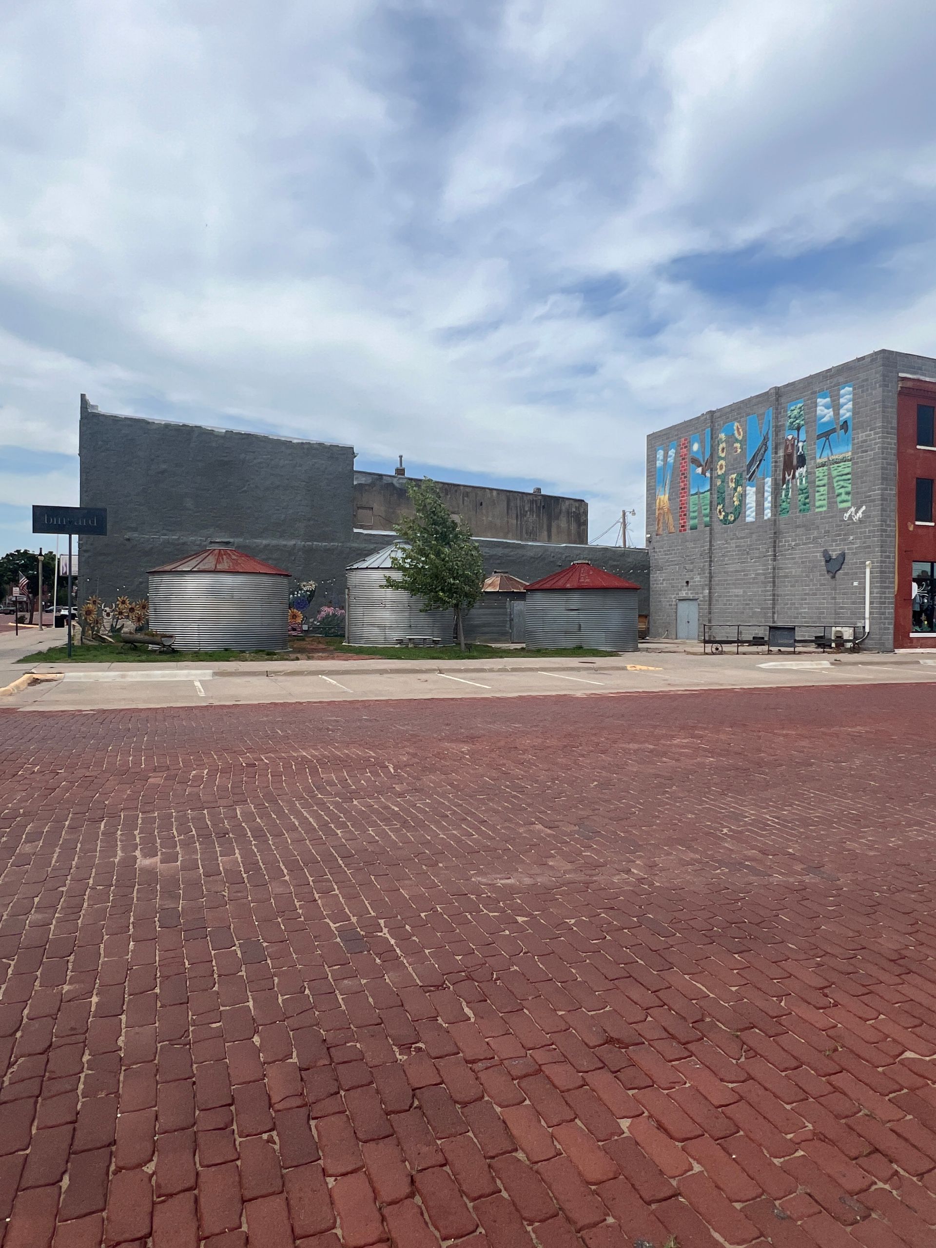Red brick plaza with modern buildings, murals, and sculptural planters under a cloudy sky