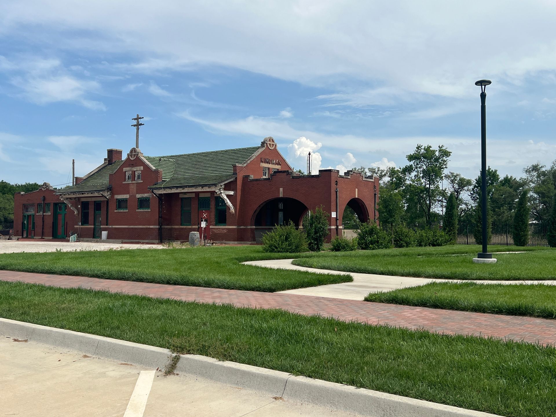 Red brick building with arched entrance and green roof beside a grassy park path under a blue sky