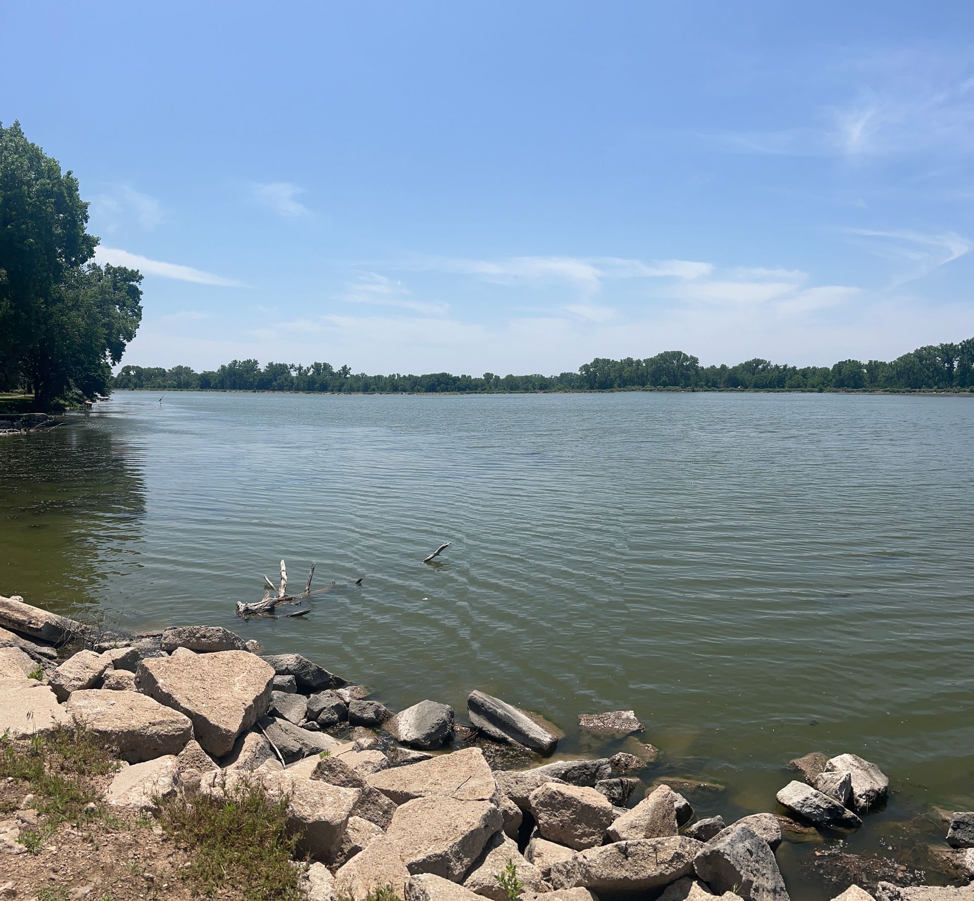 Rocky shoreline beside a calm lake under a clear blue sky, with trees lining the far shore.