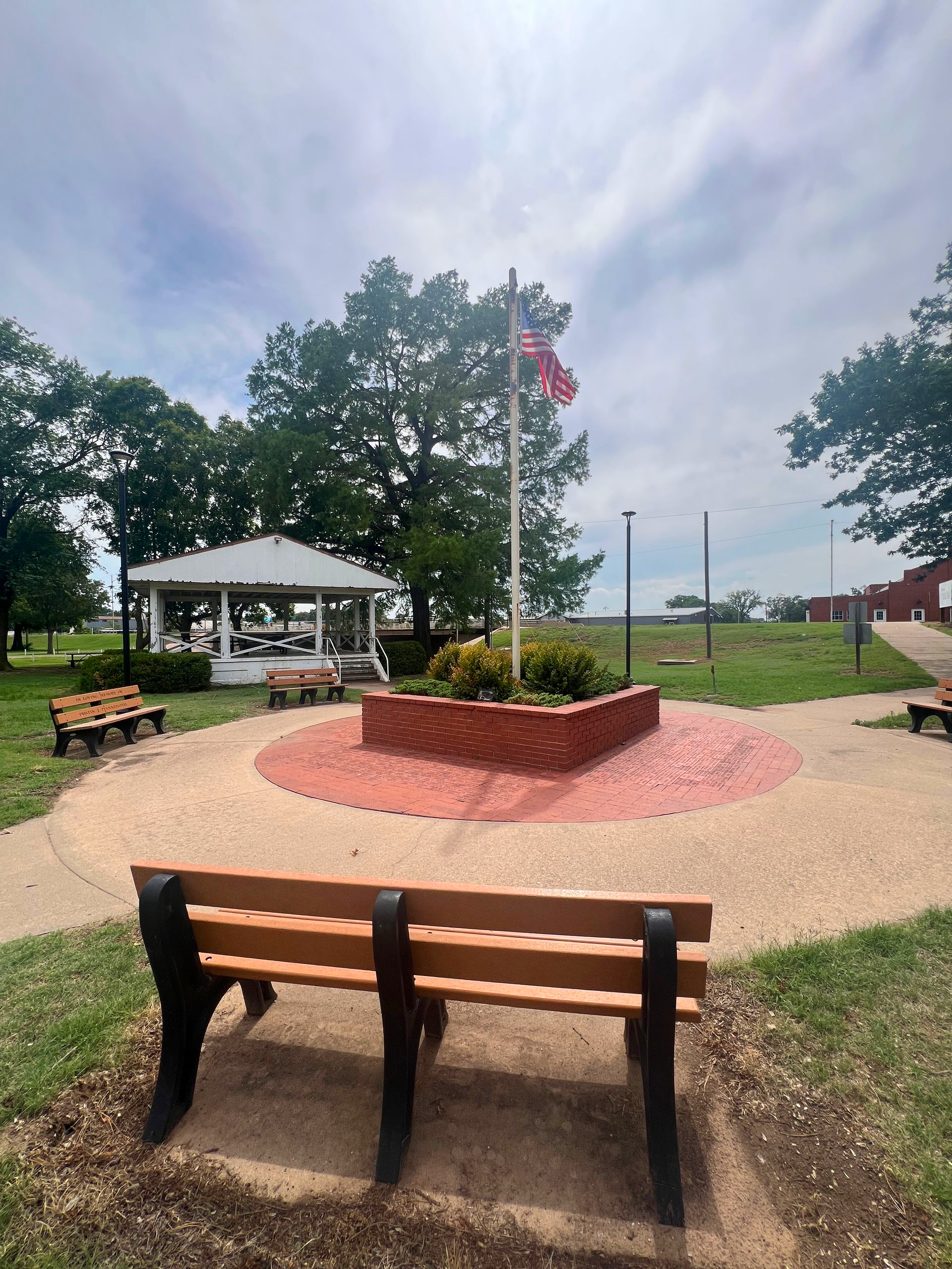 Park bench facing a flower bed and gazebo in a landscaped park under a cloudy sky