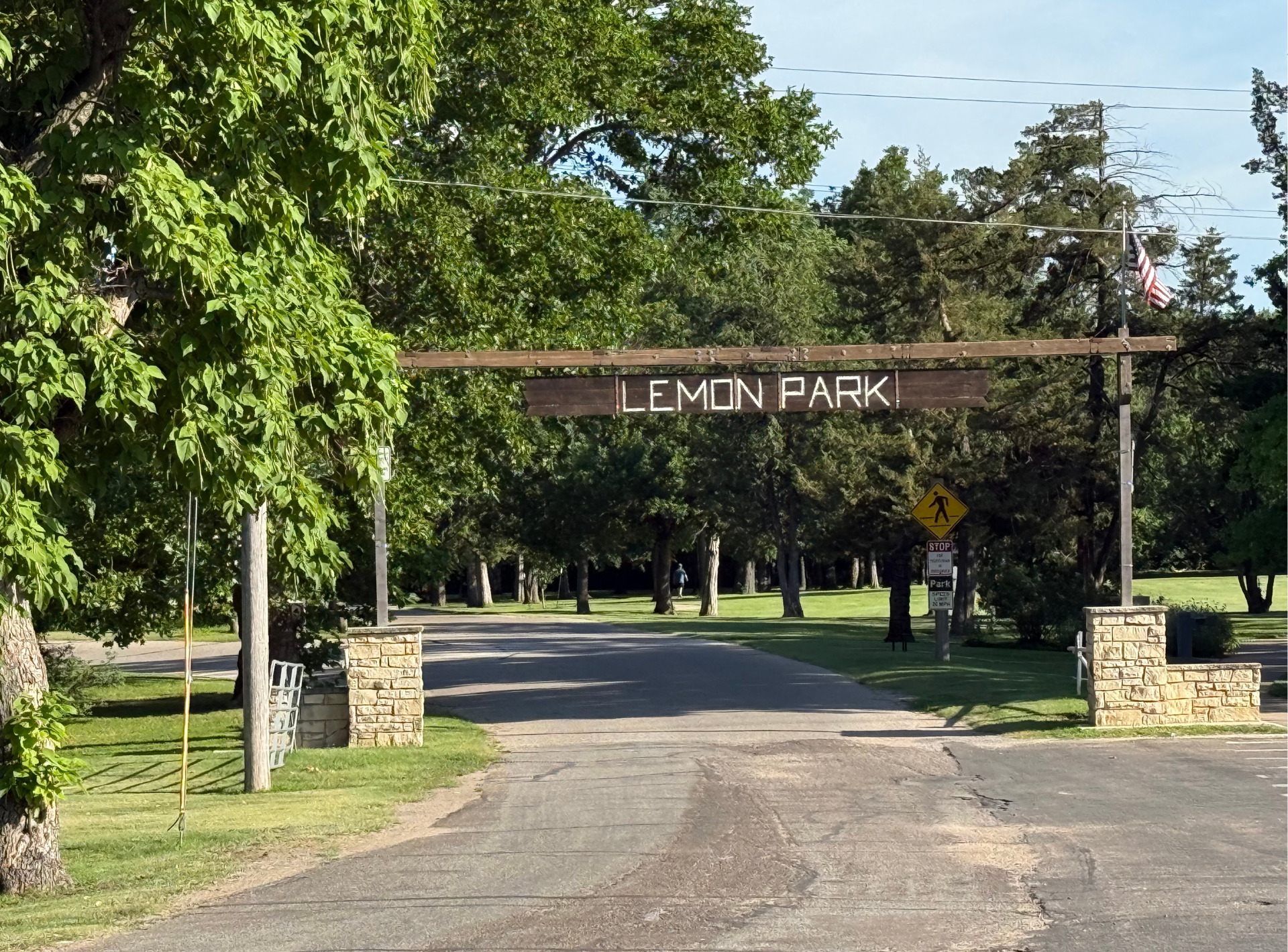 Park entrance with a wooden arch sign, stone pillars, and a paved road lined by trees.