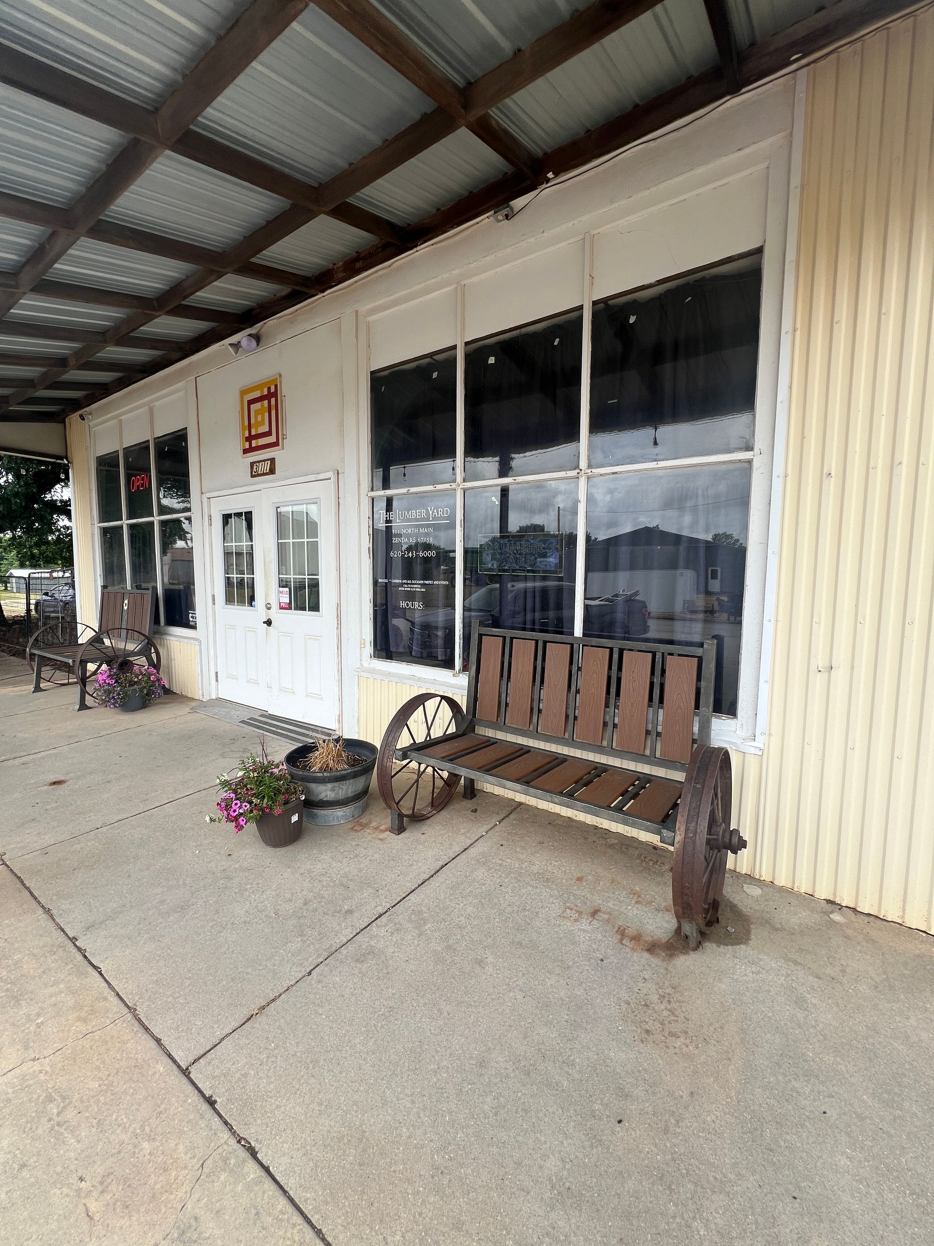 Covered storefront with windows, wooden bench, and potted flowers on a concrete sidewalk.