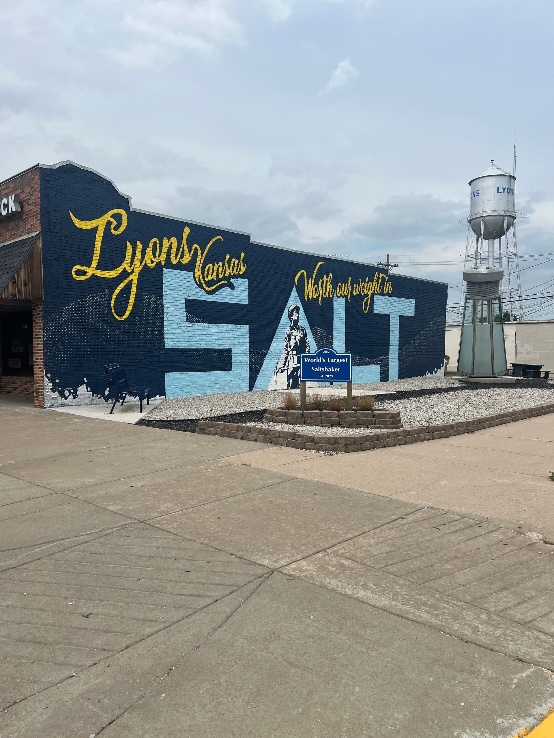 Mural on brick building with bold blue and yellow lettering, beside a water tower under a cloudy sky.