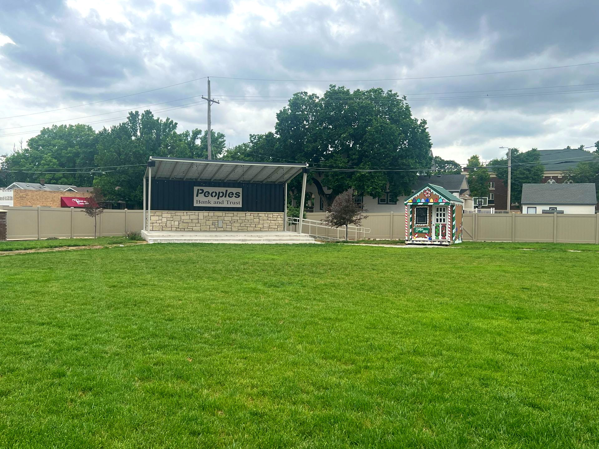 Green grassy field facing a small outdoor stage with a canopy and a colorful play structure under cloudy skies