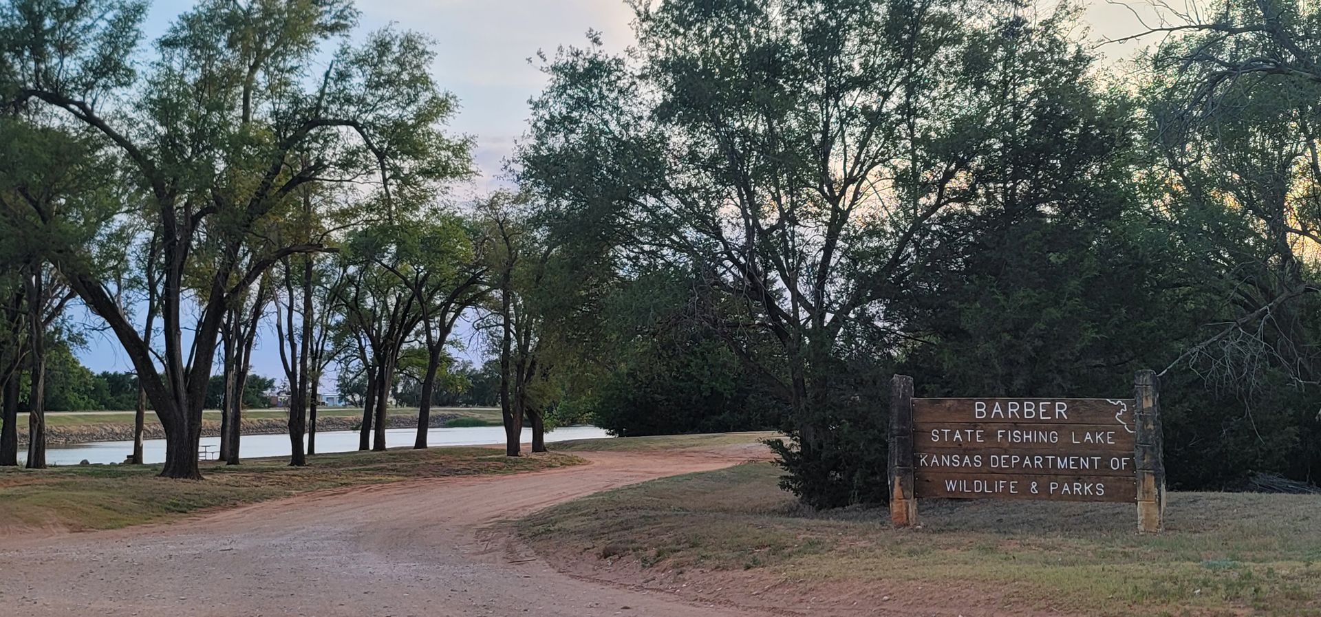 Wooded lakeside park entrance with a dirt path and a brown sign at right