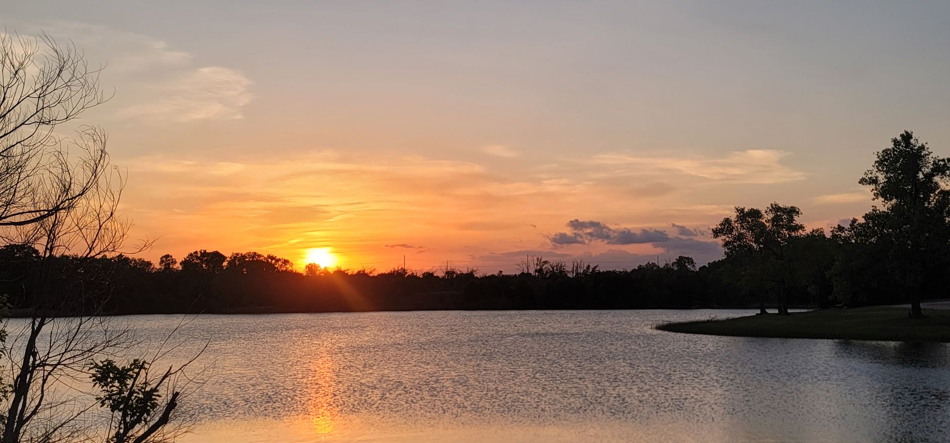 Sunset over a calm lake, with orange sky reflecting on the water and dark trees along the shore